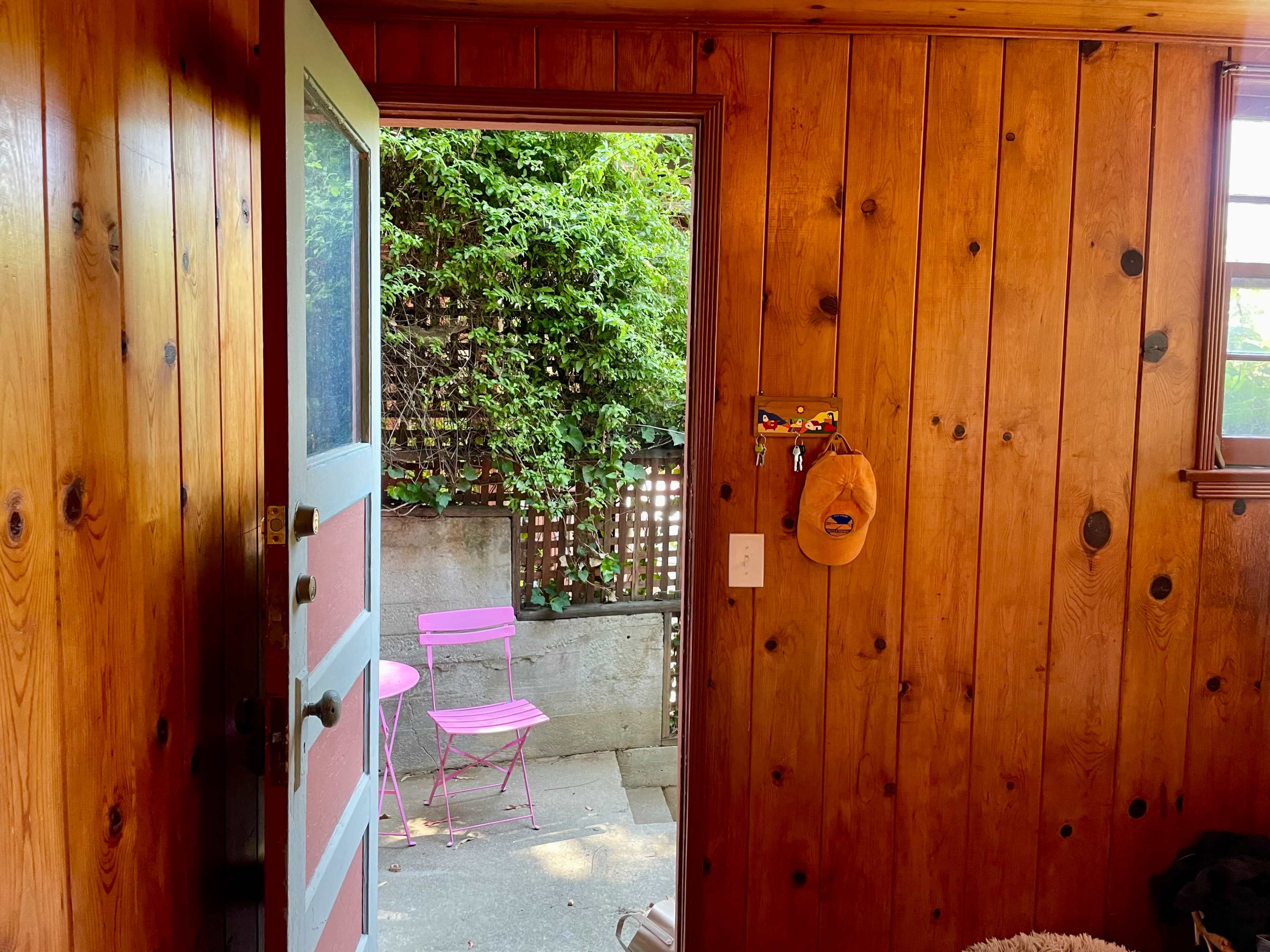 A wooden door opens to a small outdoor area with two pink chairs beside a wall covered in greenery.