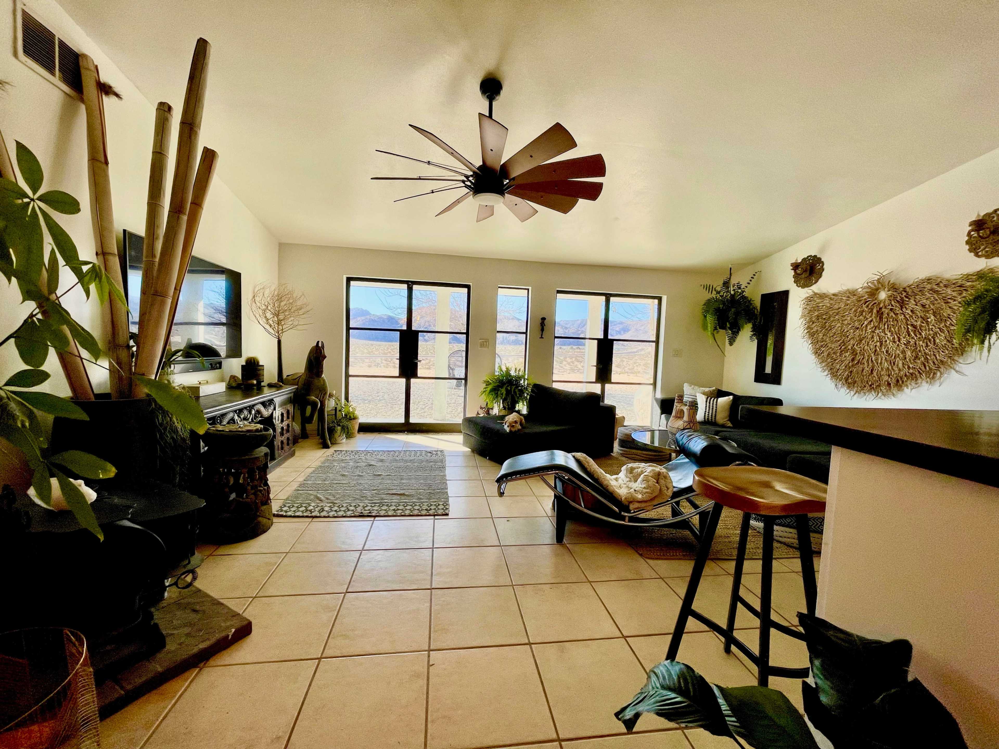 The image displays a spacious living room featuring large windows with mountain views, a ceiling fan, and various indoor plants.