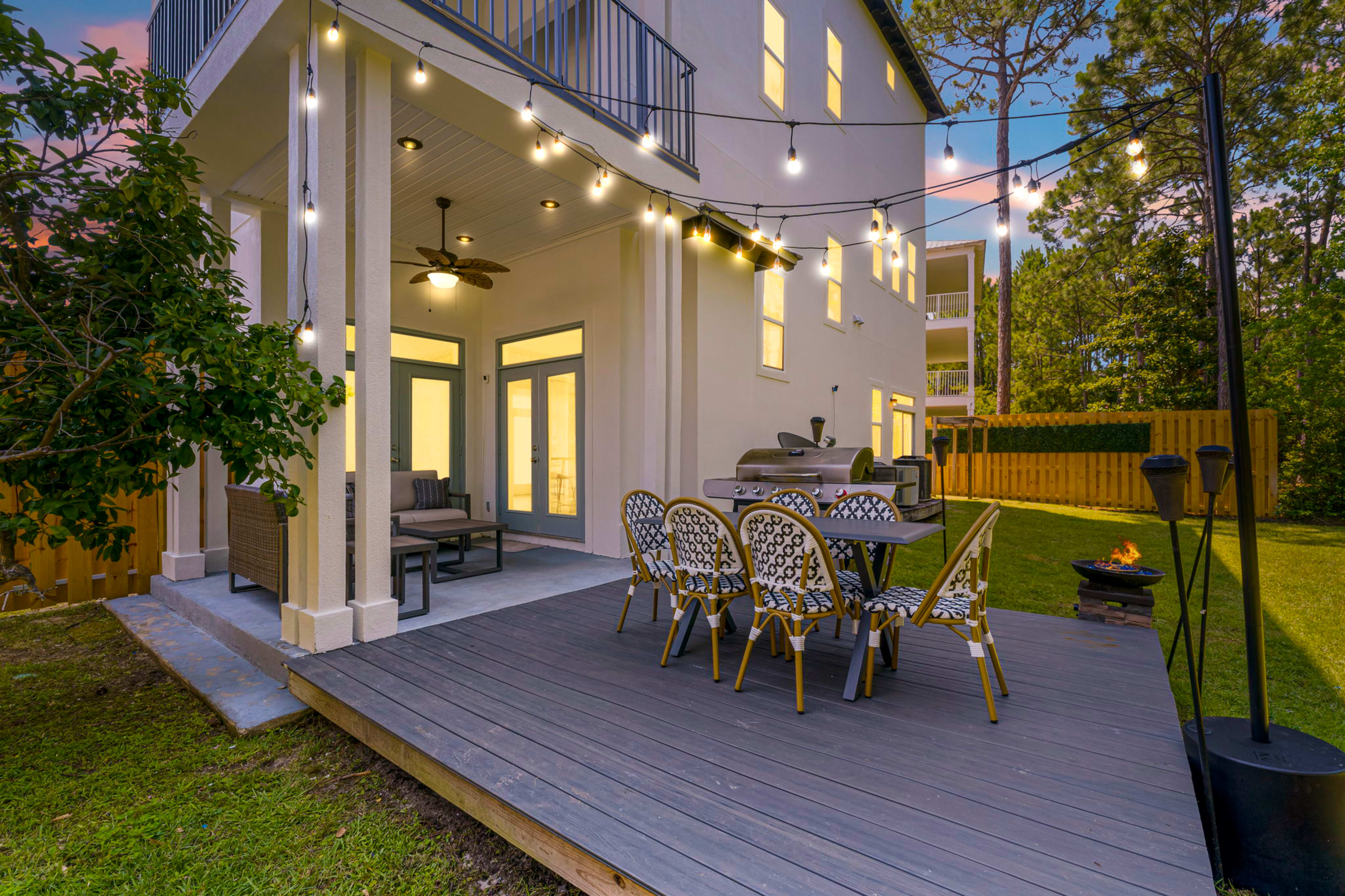 A backyard patio with string lights, a dining table with chairs, a grill, and a fire pit, adjacent to a two-story house.