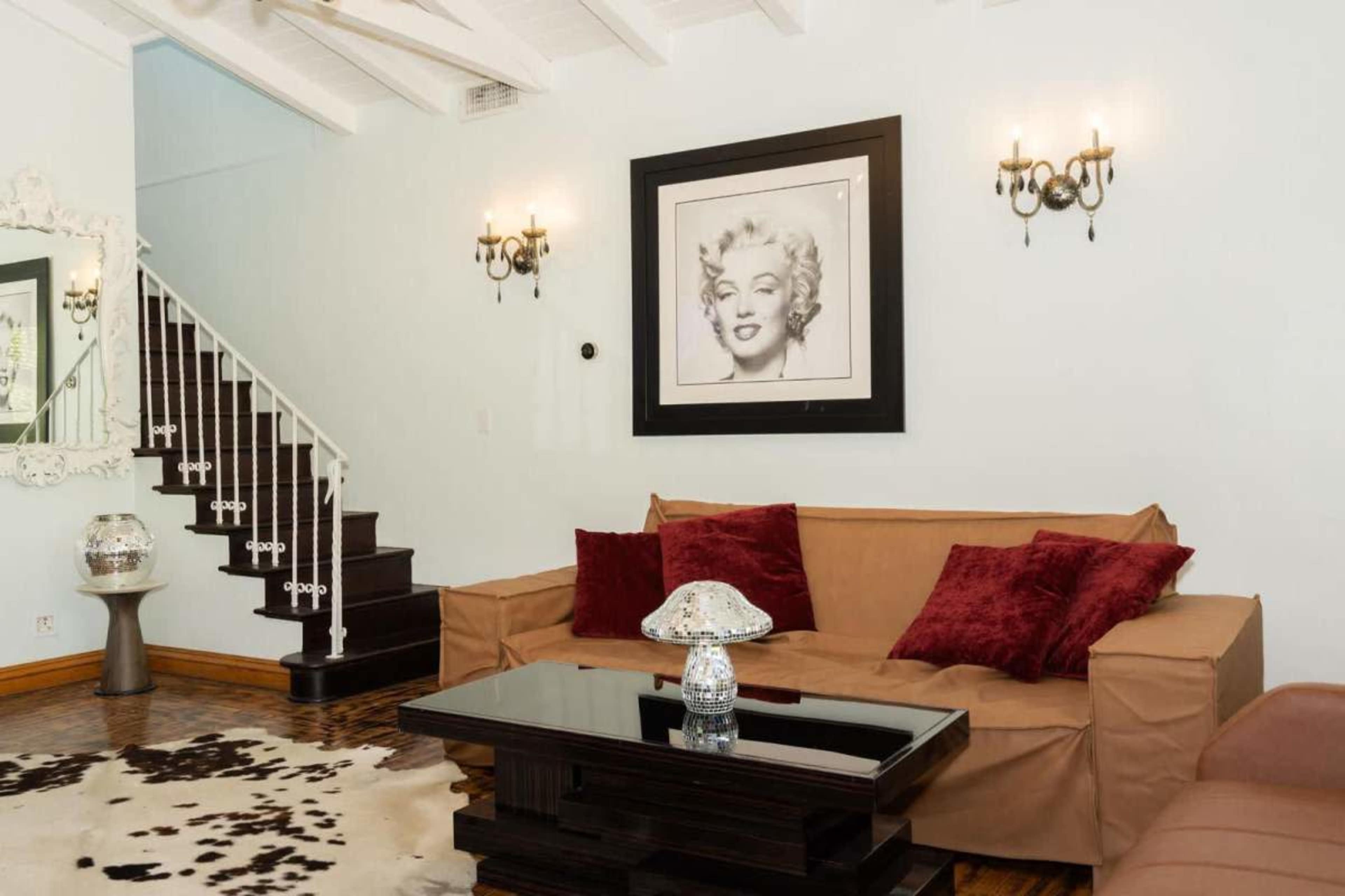The image depicts a living room featuring a beige sofa with red pillows, a glass coffee table, and a staircase leading to an upper level, accented by a large framed portrait of a woman on the wall.