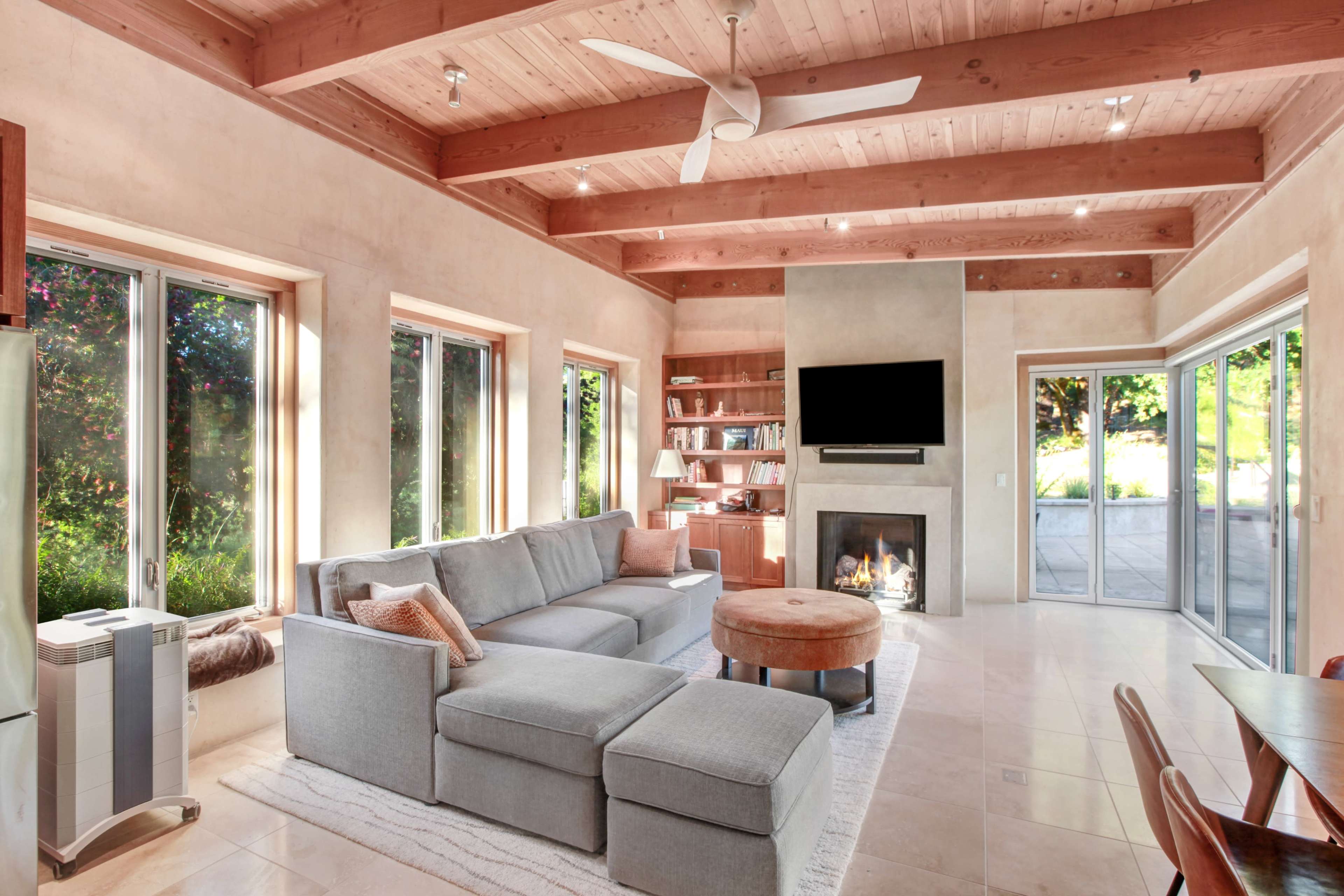A modern living room featuring a gray sectional sofa, a round coffee table, a fireplace, and large windows that overlook a landscaped outdoor area.