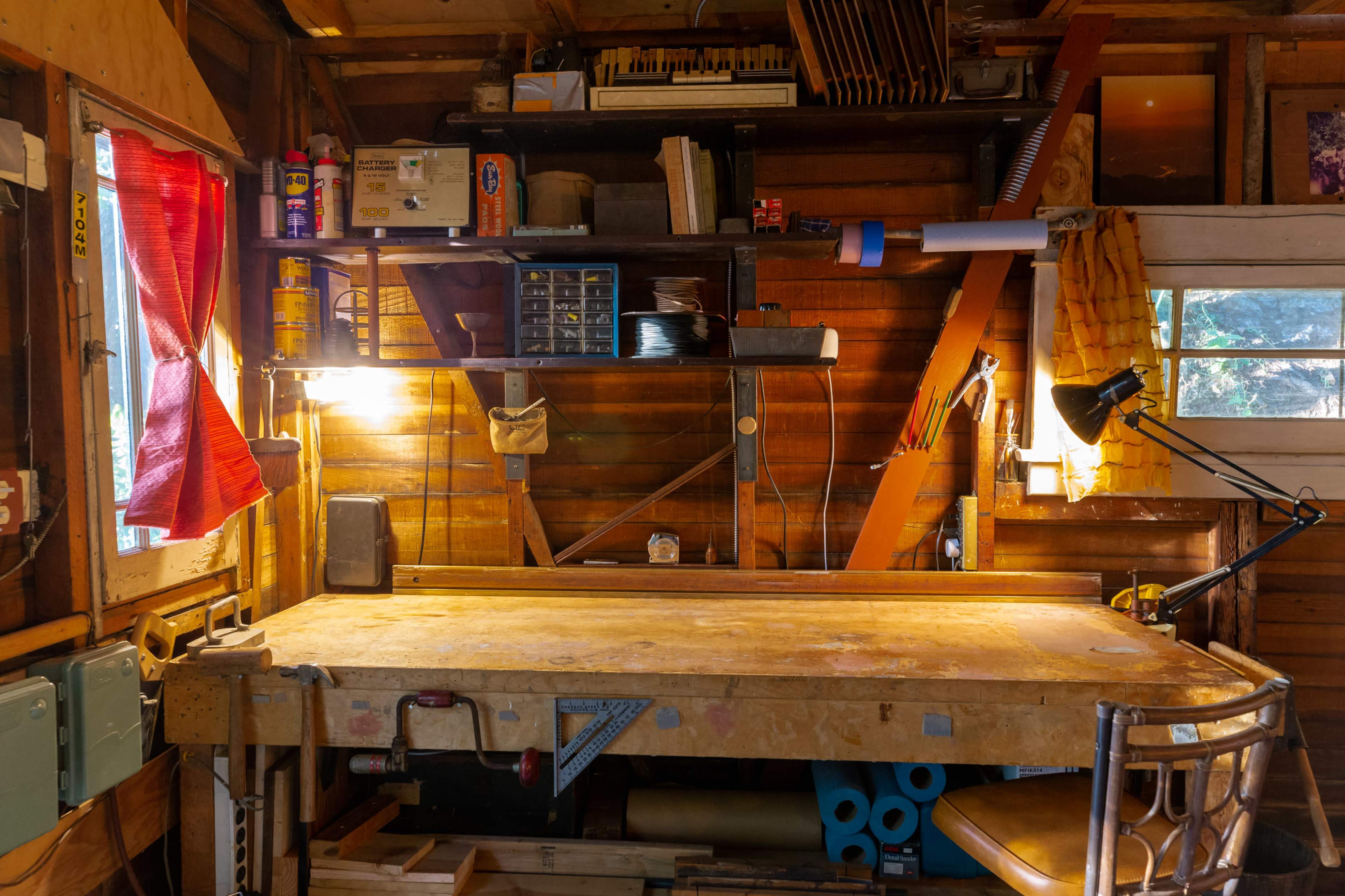 A wooden workbench is illuminated by lamps in a cluttered workshop featuring shelves filled with tools and supplies.