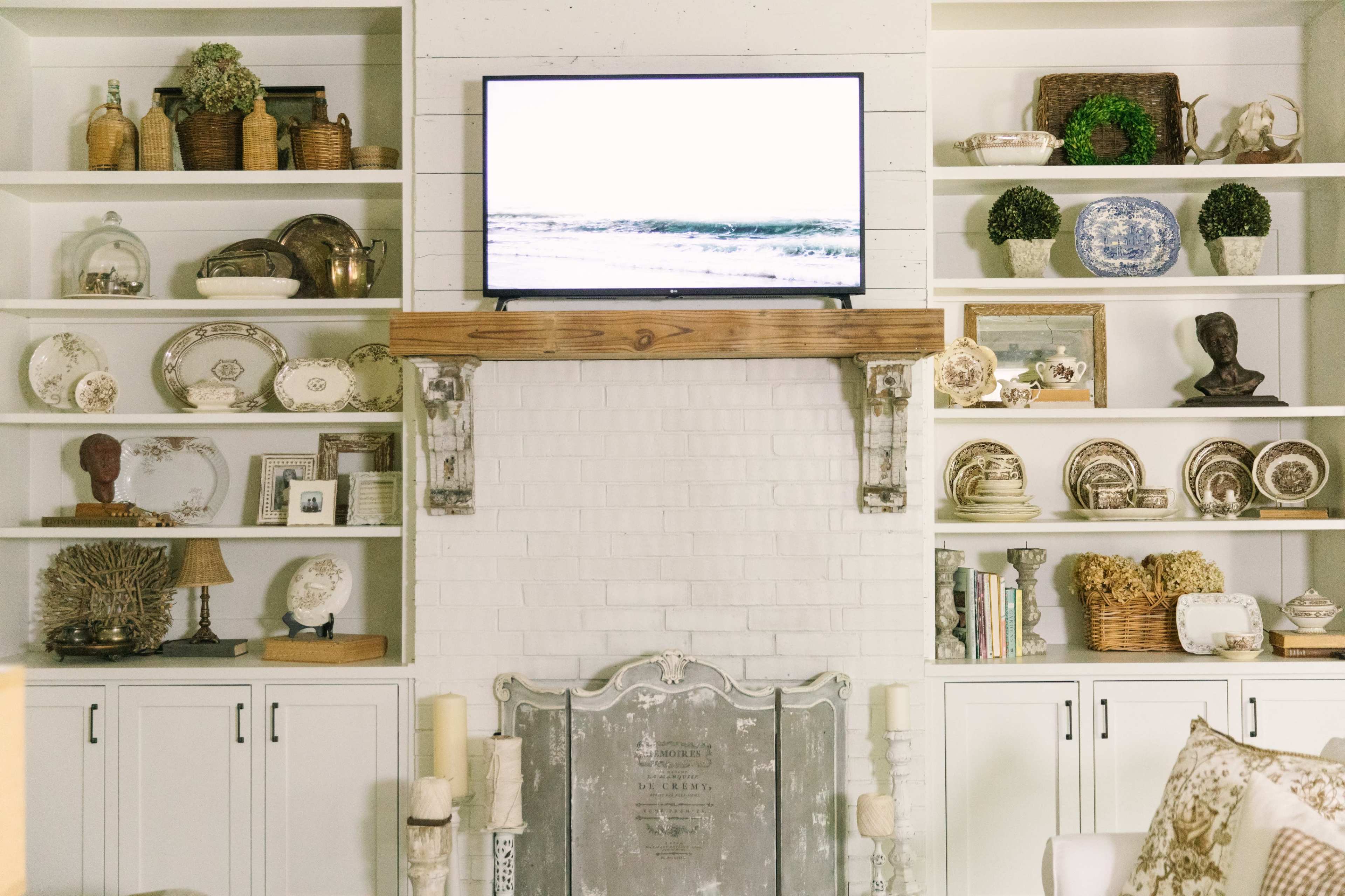 A living room features a mounted television above a wooden mantel, surrounded by decorative shelves filled with various decorative items and vintage-style plates.