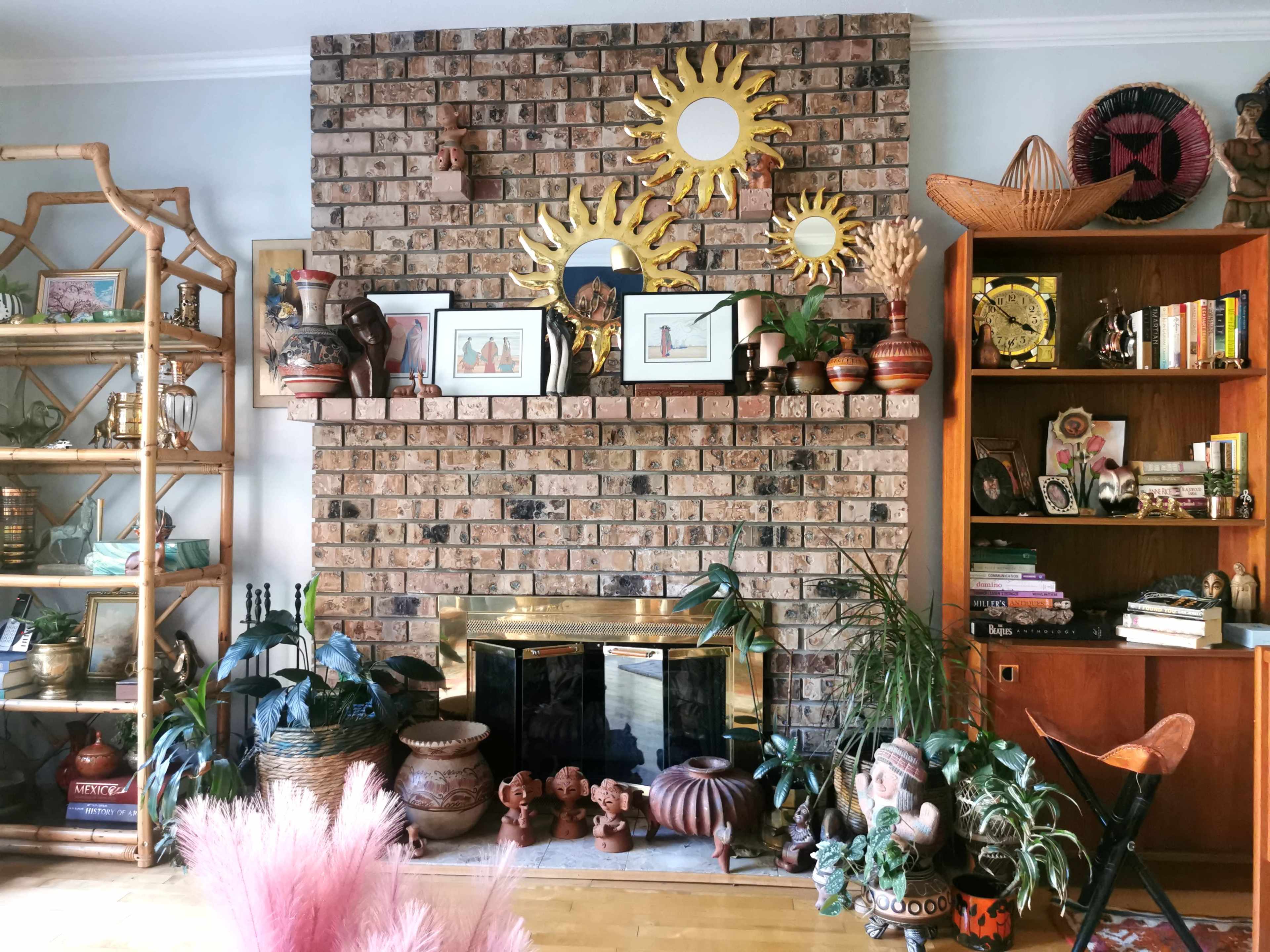 A brick fireplace is the central feature of a living room adorned with a variety of decorative items, plants, and artwork on the walls and surrounding shelves.