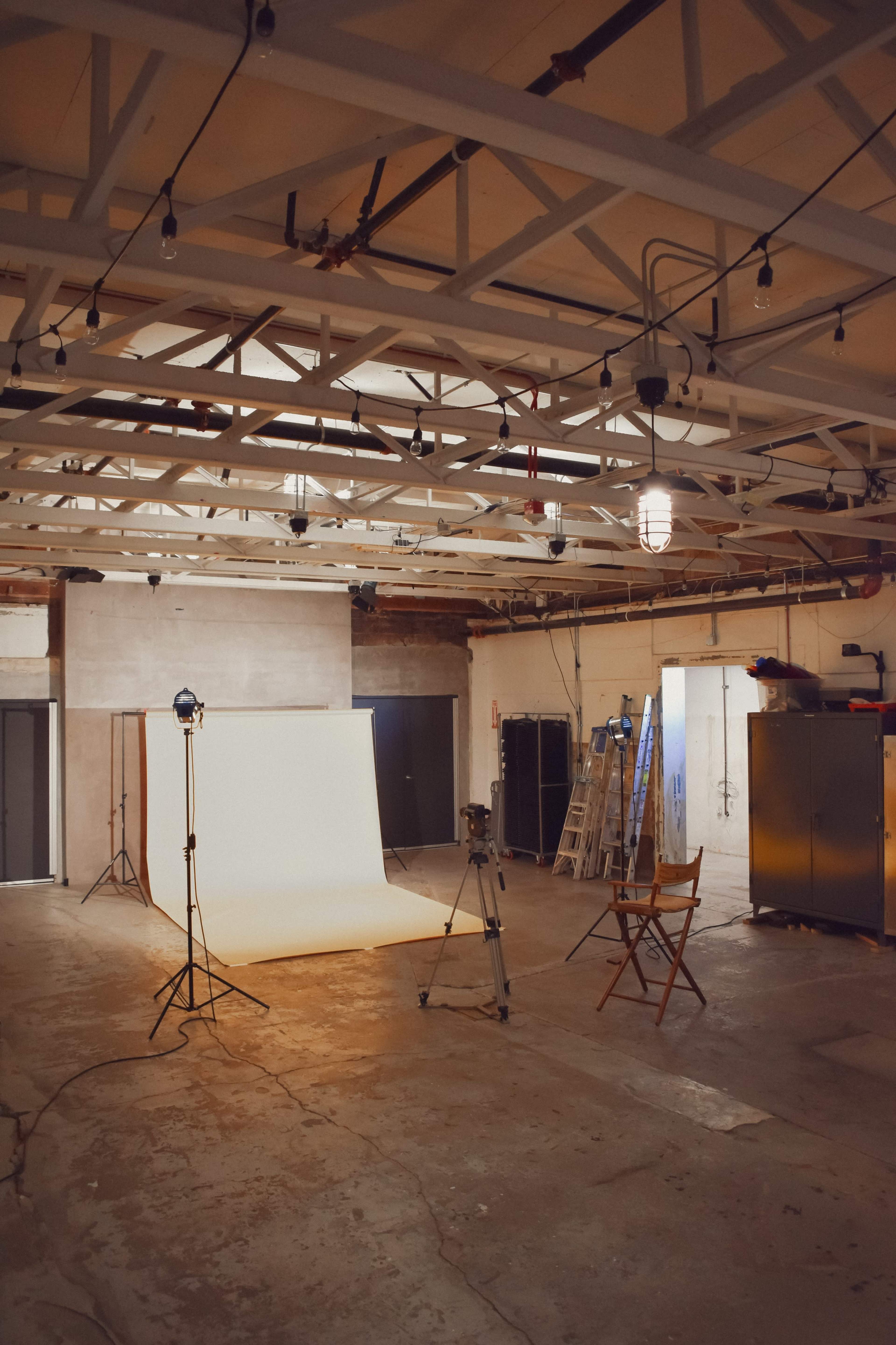 A photography studio features a light-colored backdrop, camera equipment, a director's chair, and exposed beams overhead.