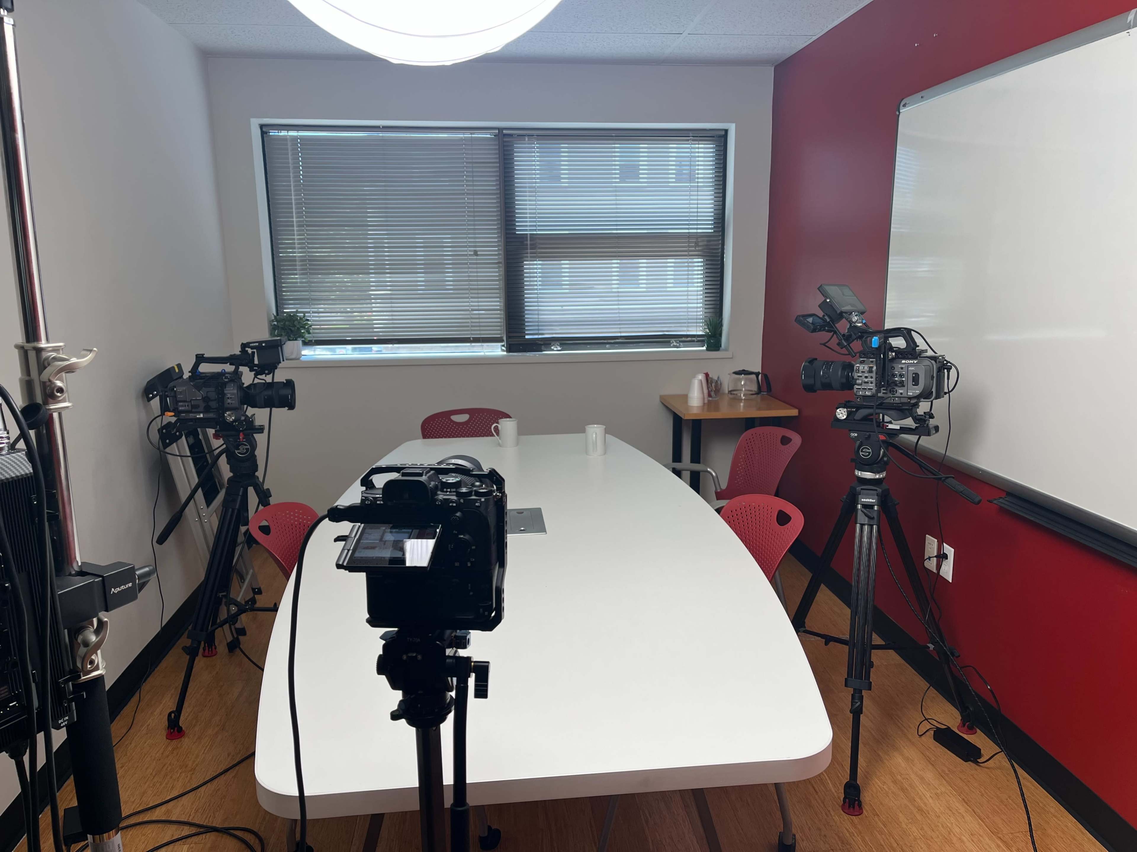 The image shows a conference room set up with a white table surrounded by red chairs, equipped with three video cameras and a whiteboard on the wall.