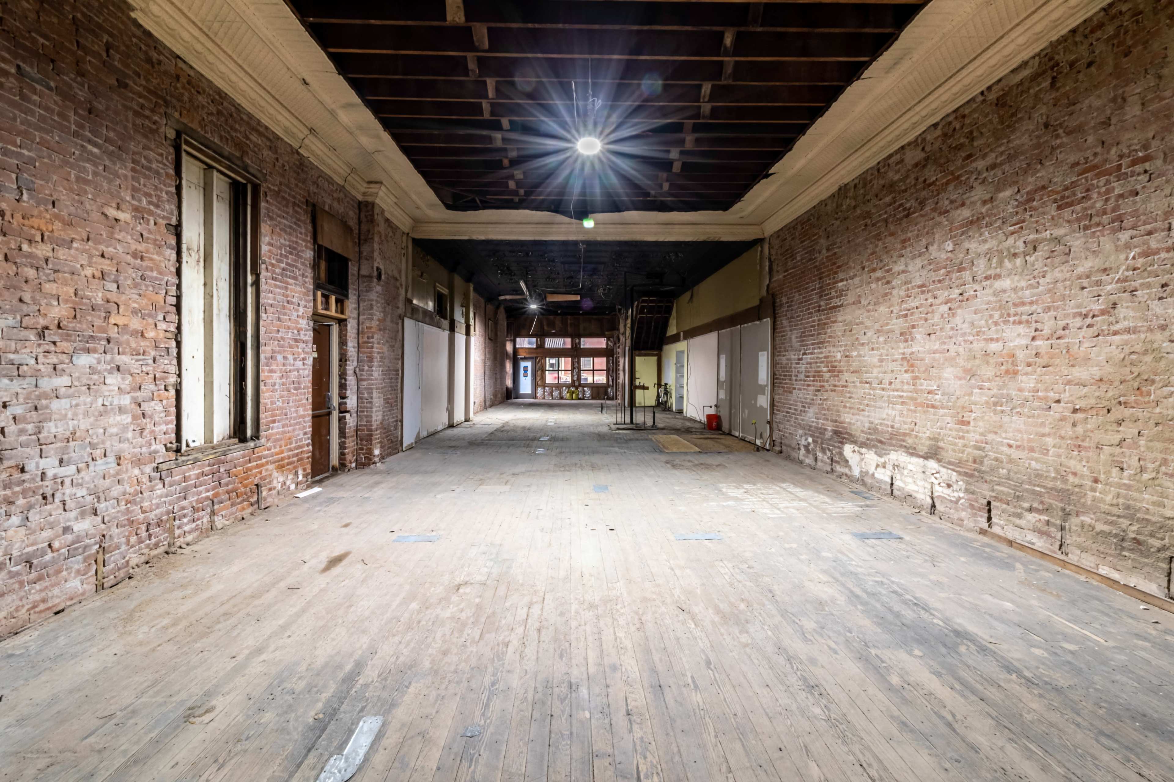 A long, empty hall with exposed brick walls and wooden flooring, featuring overhead lighting and a distant doorway.
