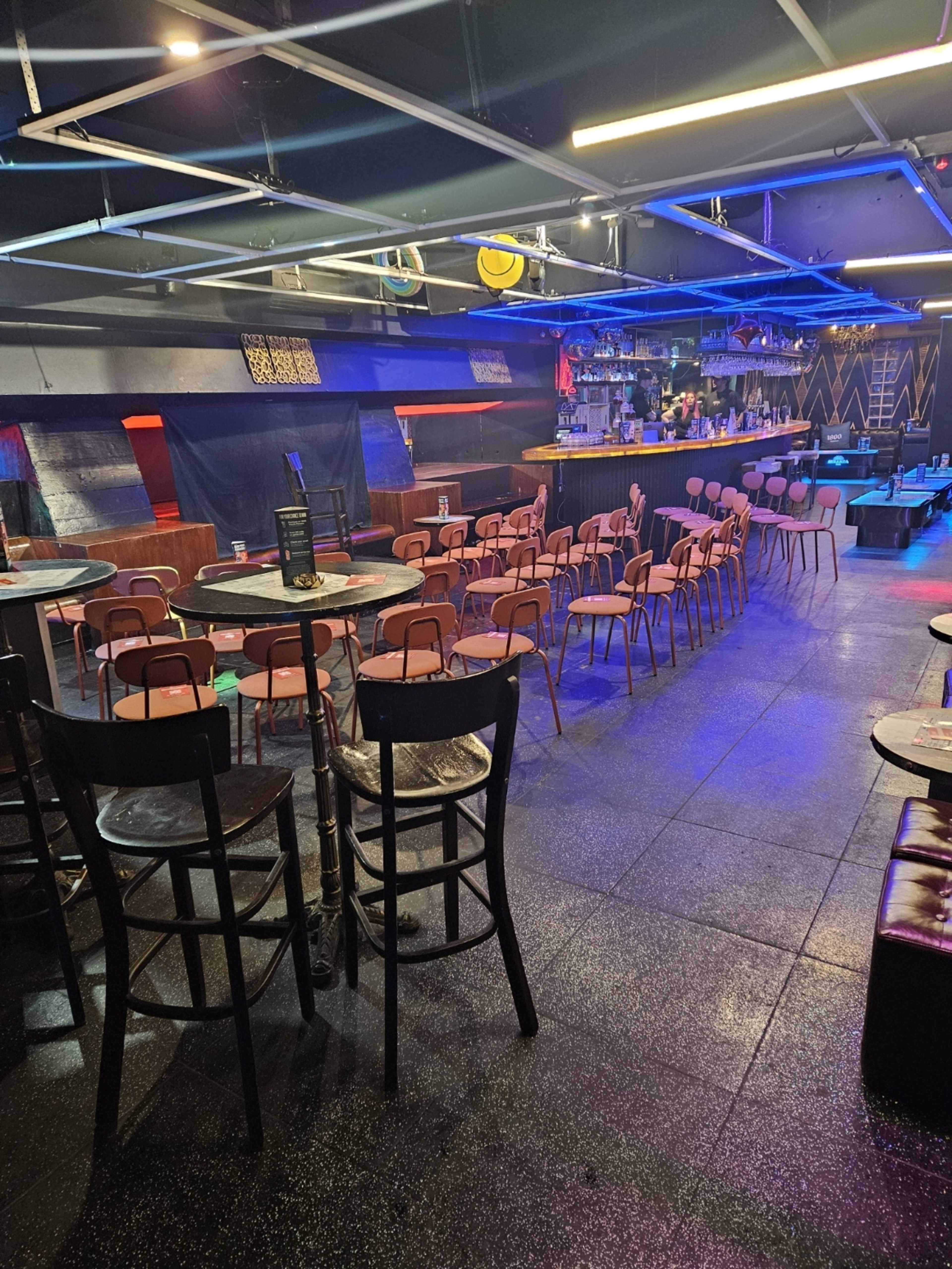The image shows a dimly lit bar interior with chairs and tables arranged in rows, a long bar counter in the background, and various colorful lights illuminating the space.