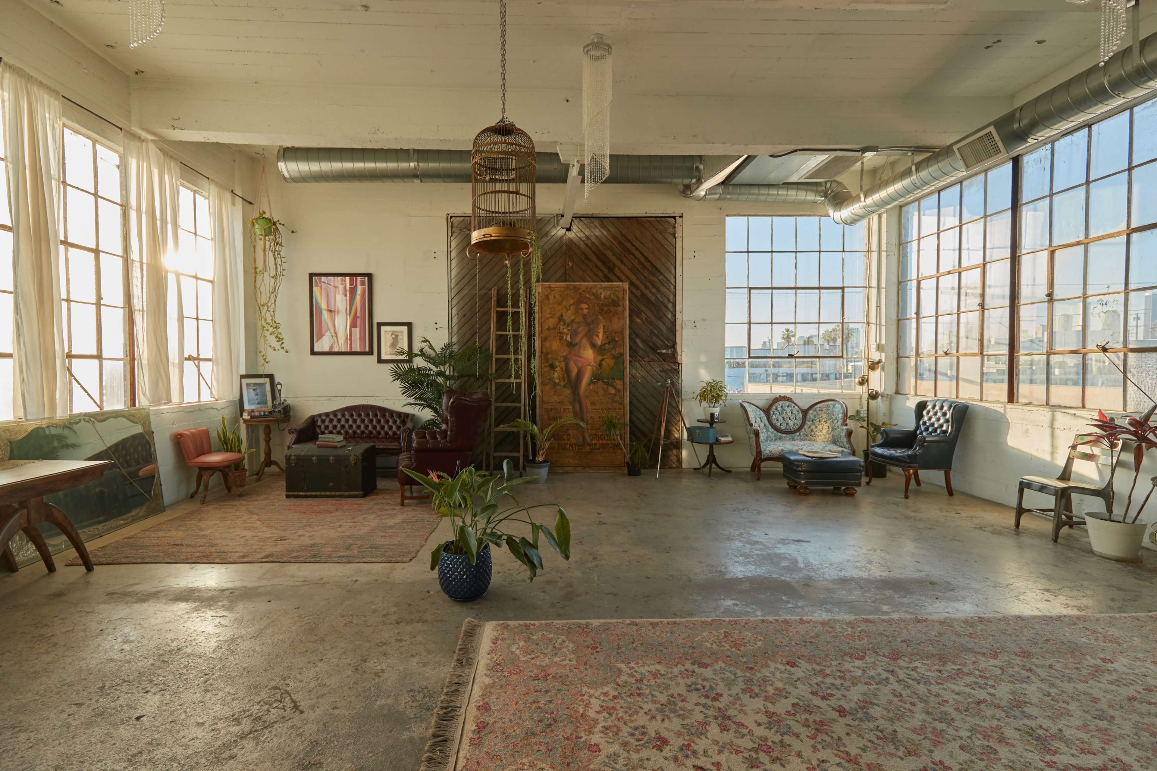 The interior of a spacious loft features large windows, a mix of seating areas, artwork, and plants on a concrete floor.