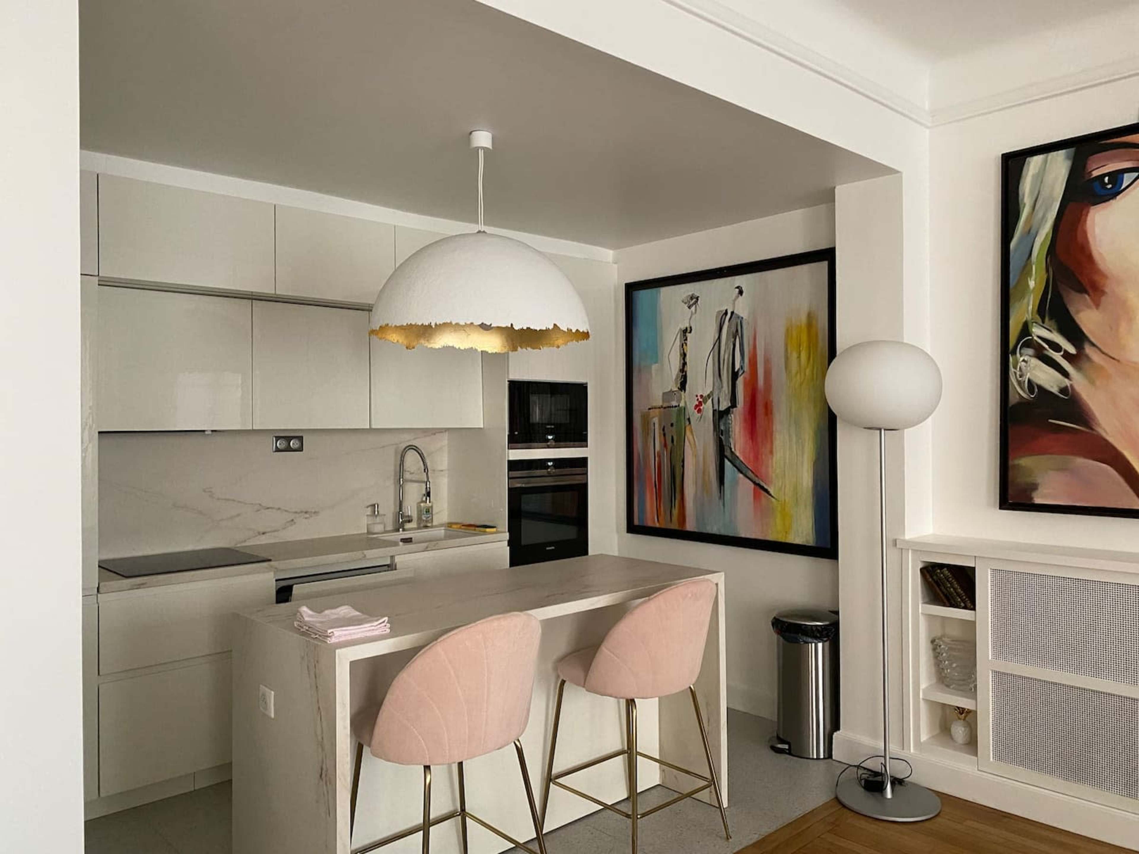 The image shows a modern kitchen area featuring a marble countertop island with two pink bar stools, a pendant light, and framed artwork on the walls.