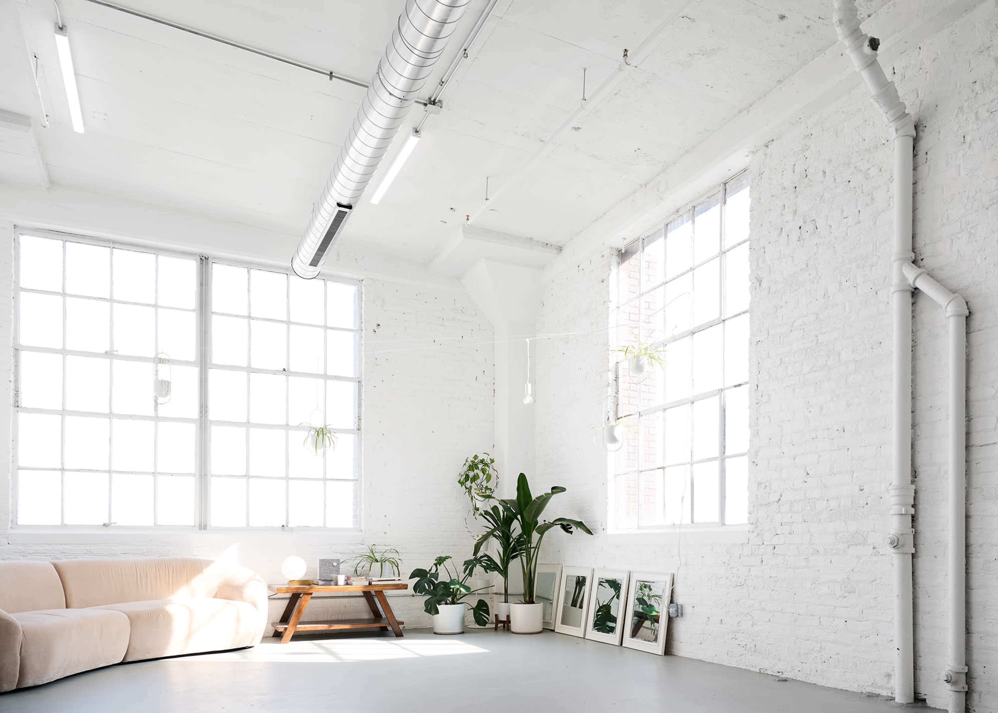 A bright, minimalist room features large windows, a light-colored sofa, and potted plants along with a wooden table and framed pictures on the floor.