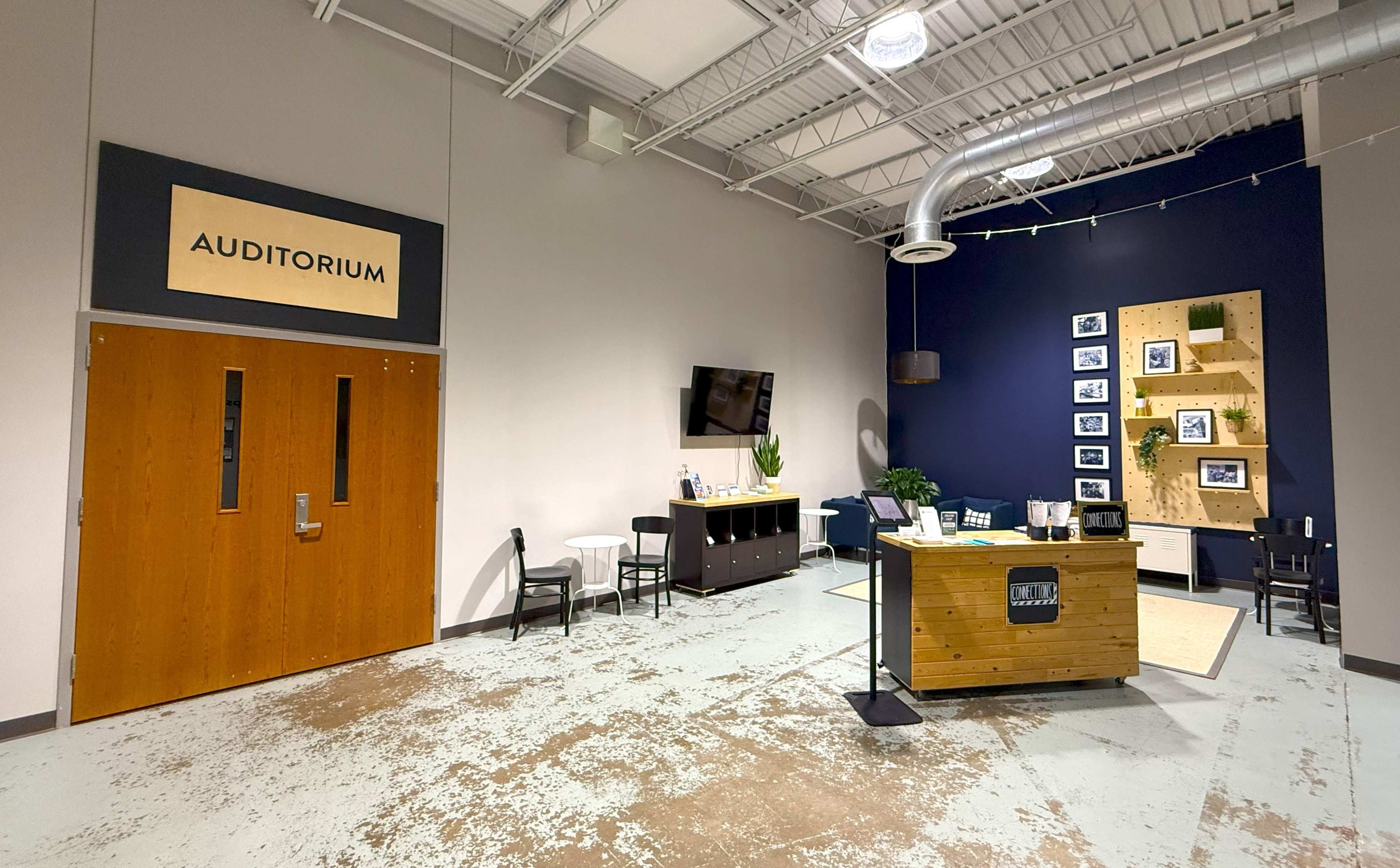 The image shows a modern lobby area with a large sign reading "AUDITORIUM," wooden doors, a reception desk, chairs, a television, and a decorative wall with framed photos.