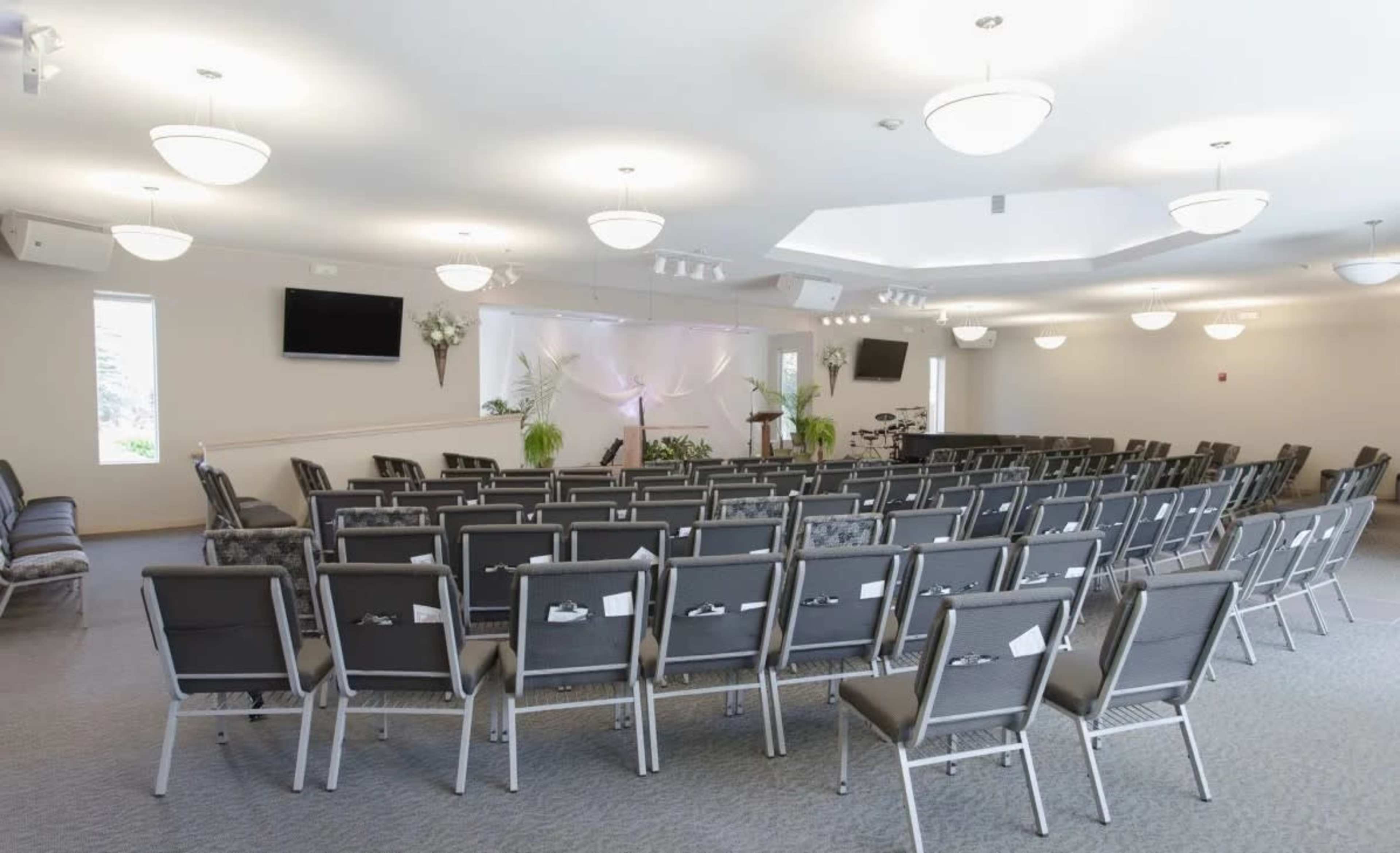 An empty auditorium with rows of metal chairs facing a podium and a decorated backdrop, flanked by plants and screens on the walls.