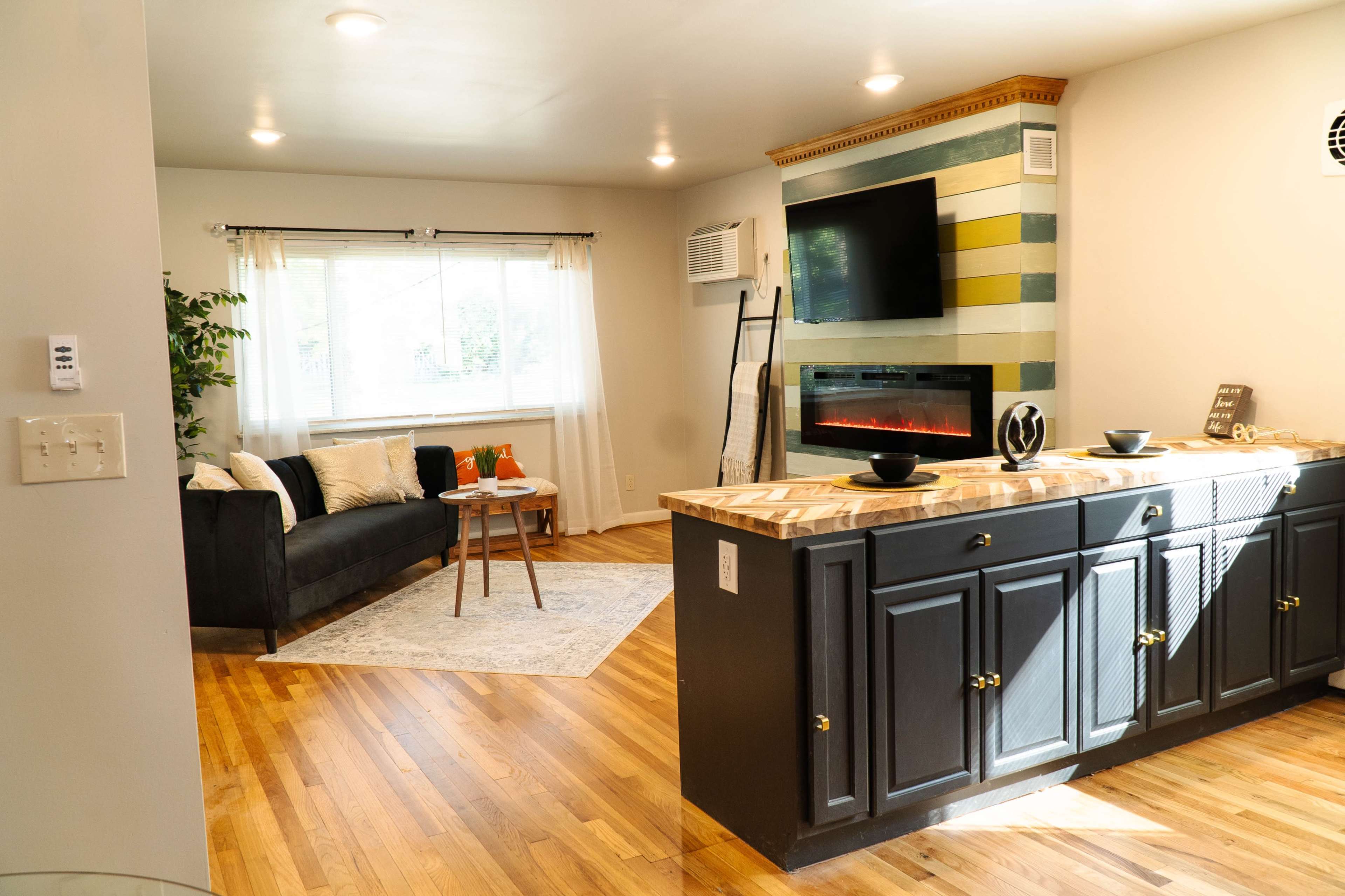 A modern living space featuring a dark sofa, an electric fireplace with a mounted TV above, and a kitchen bar with dark cabinets, all set against hardwood floors.
