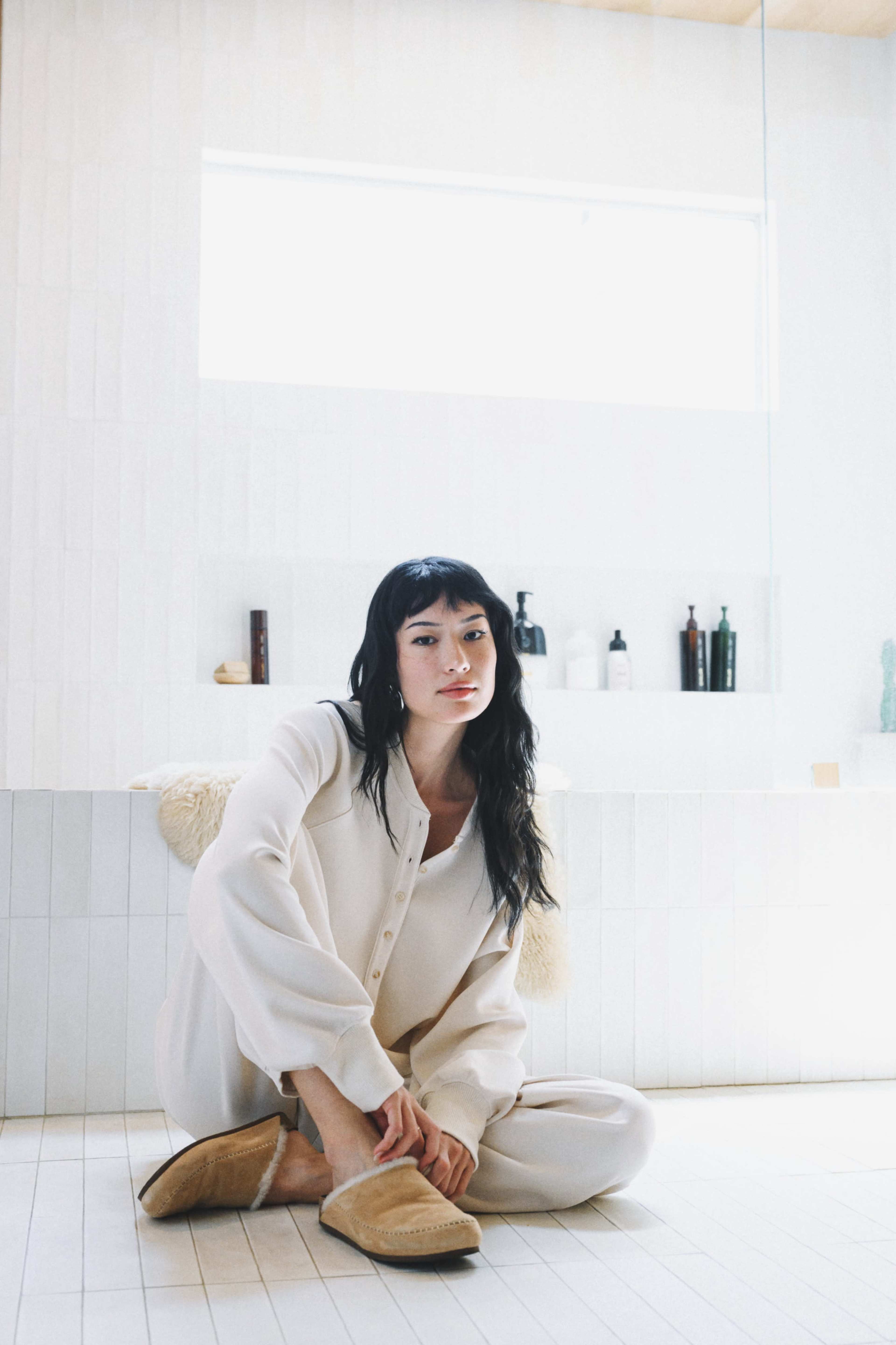 A woman sits on the floor of a bathroom, dressed in a light-colored outfit, with a variety of bottles displayed on a shelf behind her.