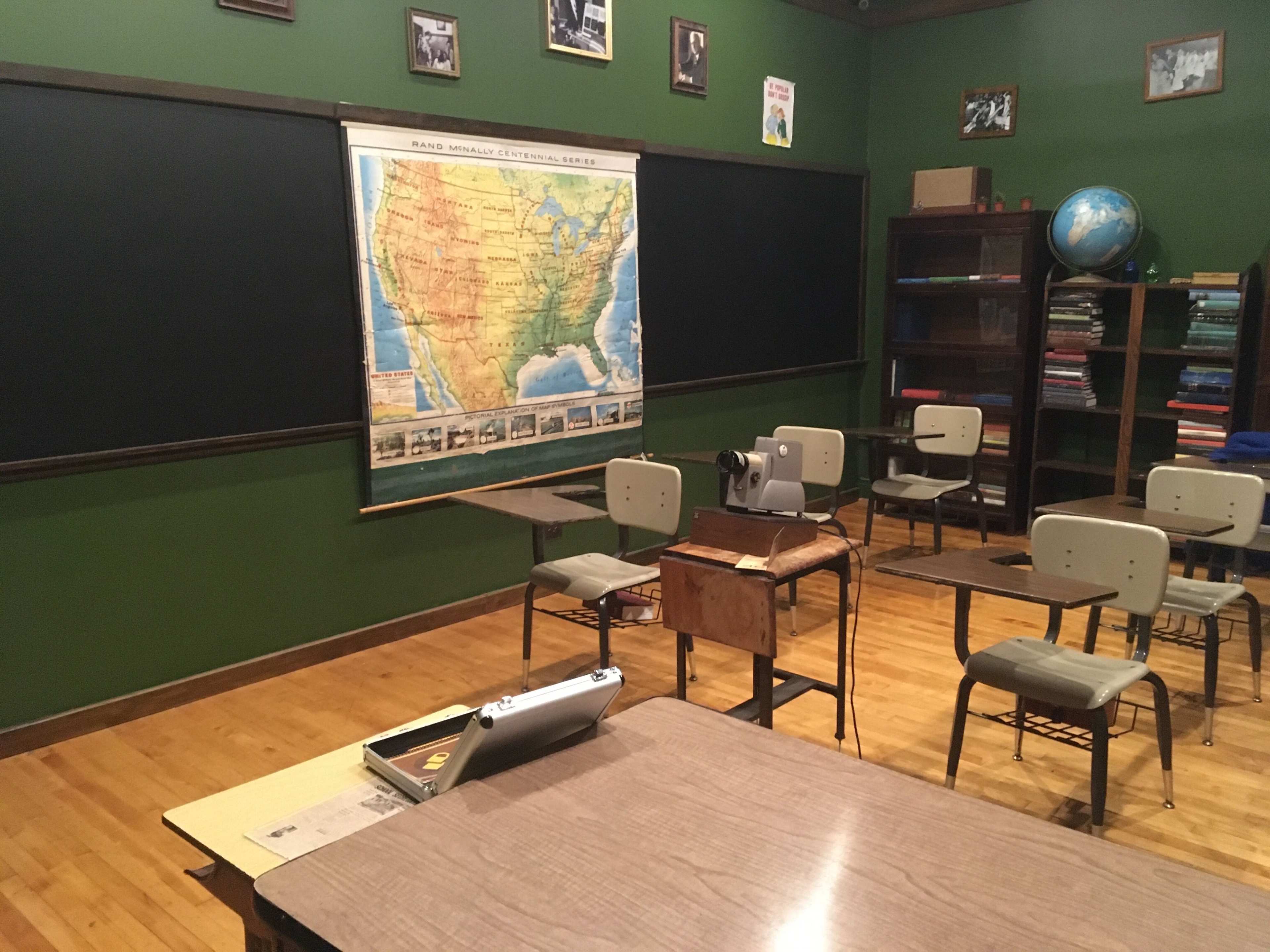 A classroom with green walls, wooden floors, desks arranged in rows, and a large map of the United States on the wall next to a globe.
