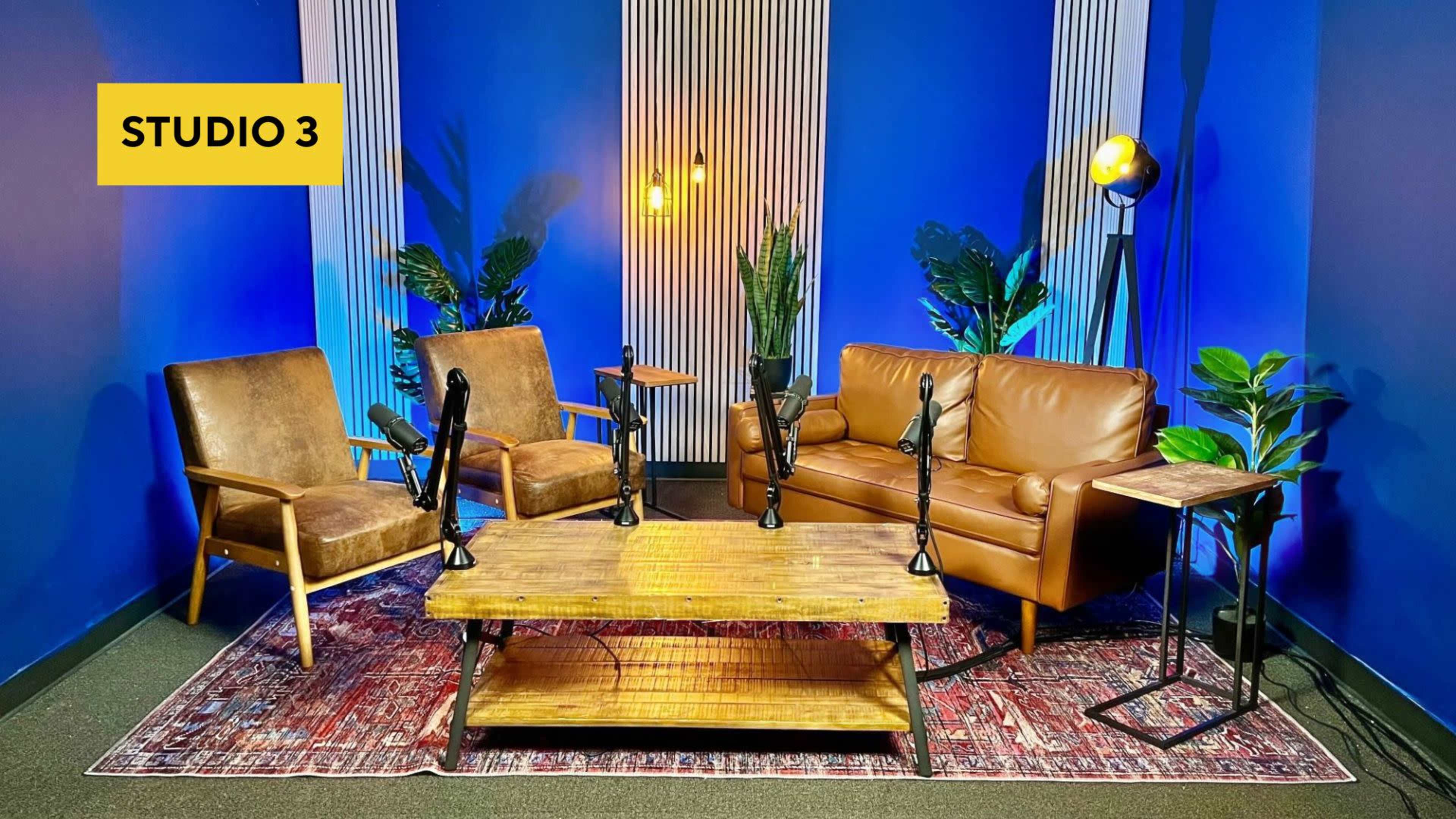 A cozy studio setup with two armchairs and a brown leather couch arranged around a wooden coffee table, surrounded by decorative plants and blue walls.