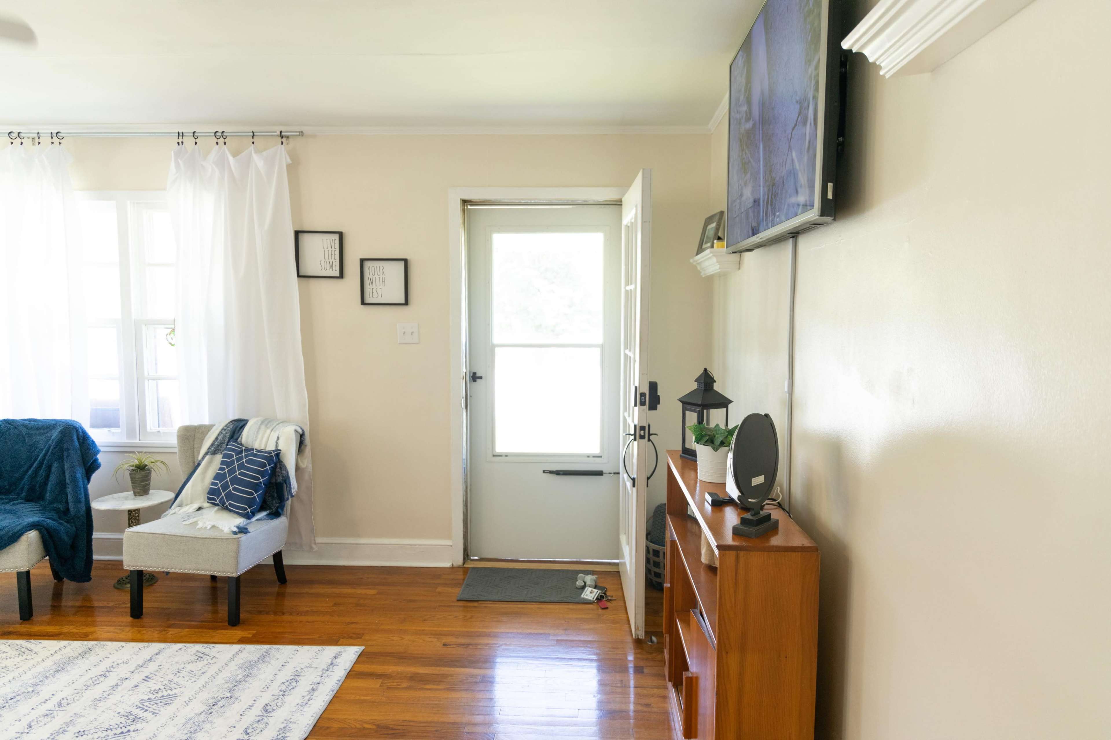 A bright living room with a door leading outside, a television mounted on the wall, and a small wooden cabinet beside a chair.