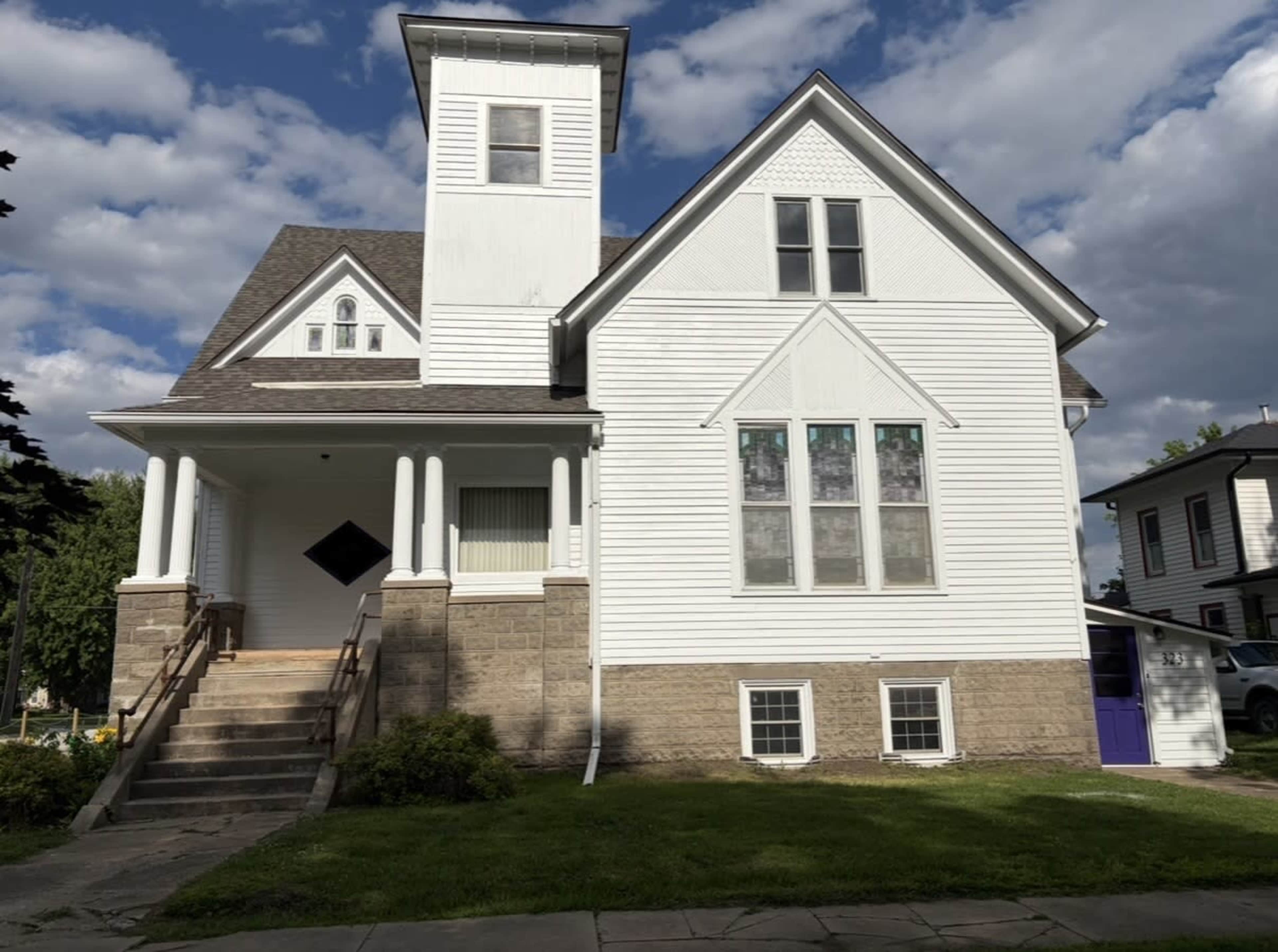 The image shows a white two-story house with a stone foundation and a prominent tower on one side, featuring large windows and a front porch with steps leading up to it.