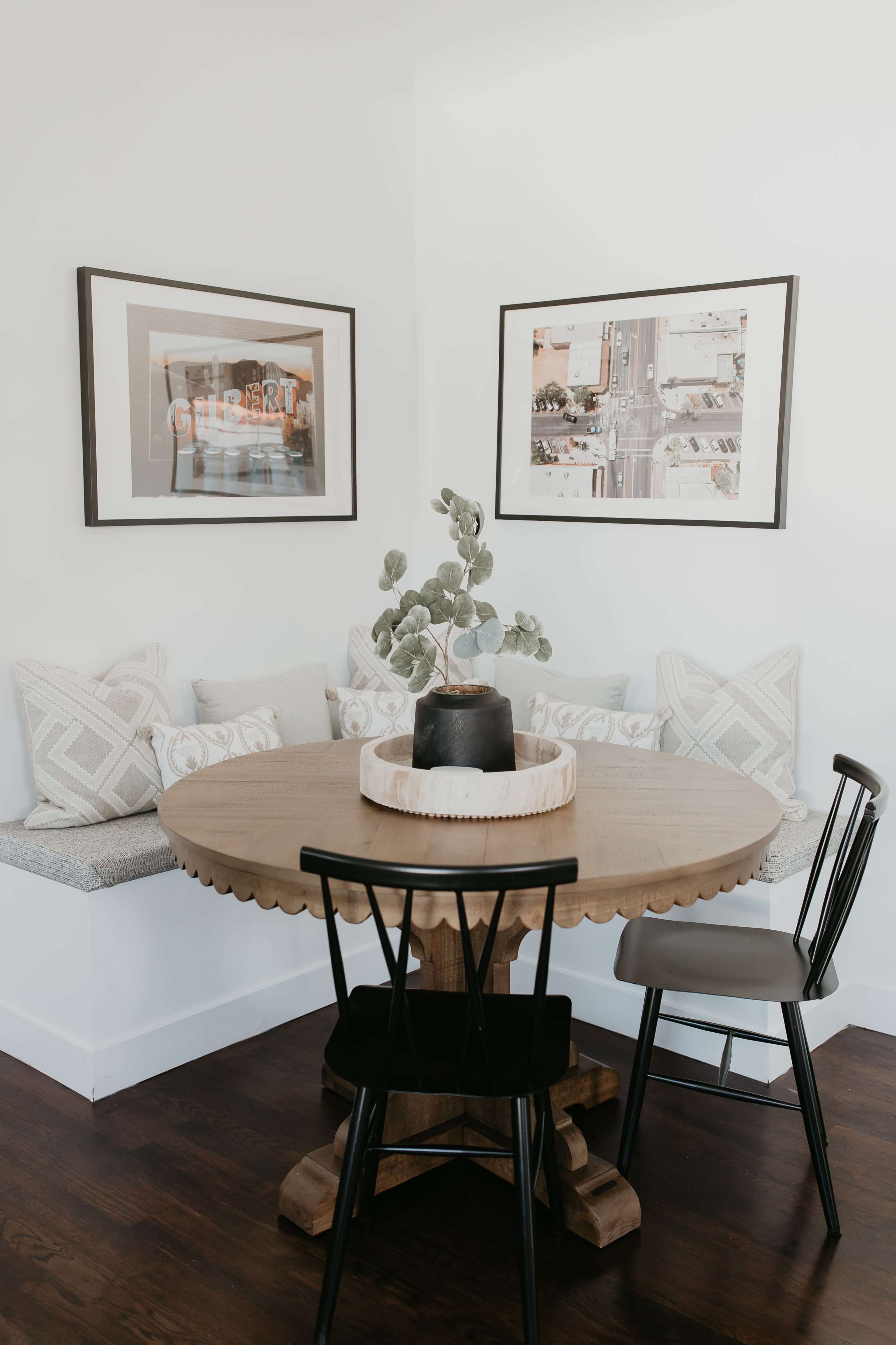 A round wooden table is set in a corner with a decorative plant and two chairs, surrounded by framed artwork on the walls.
