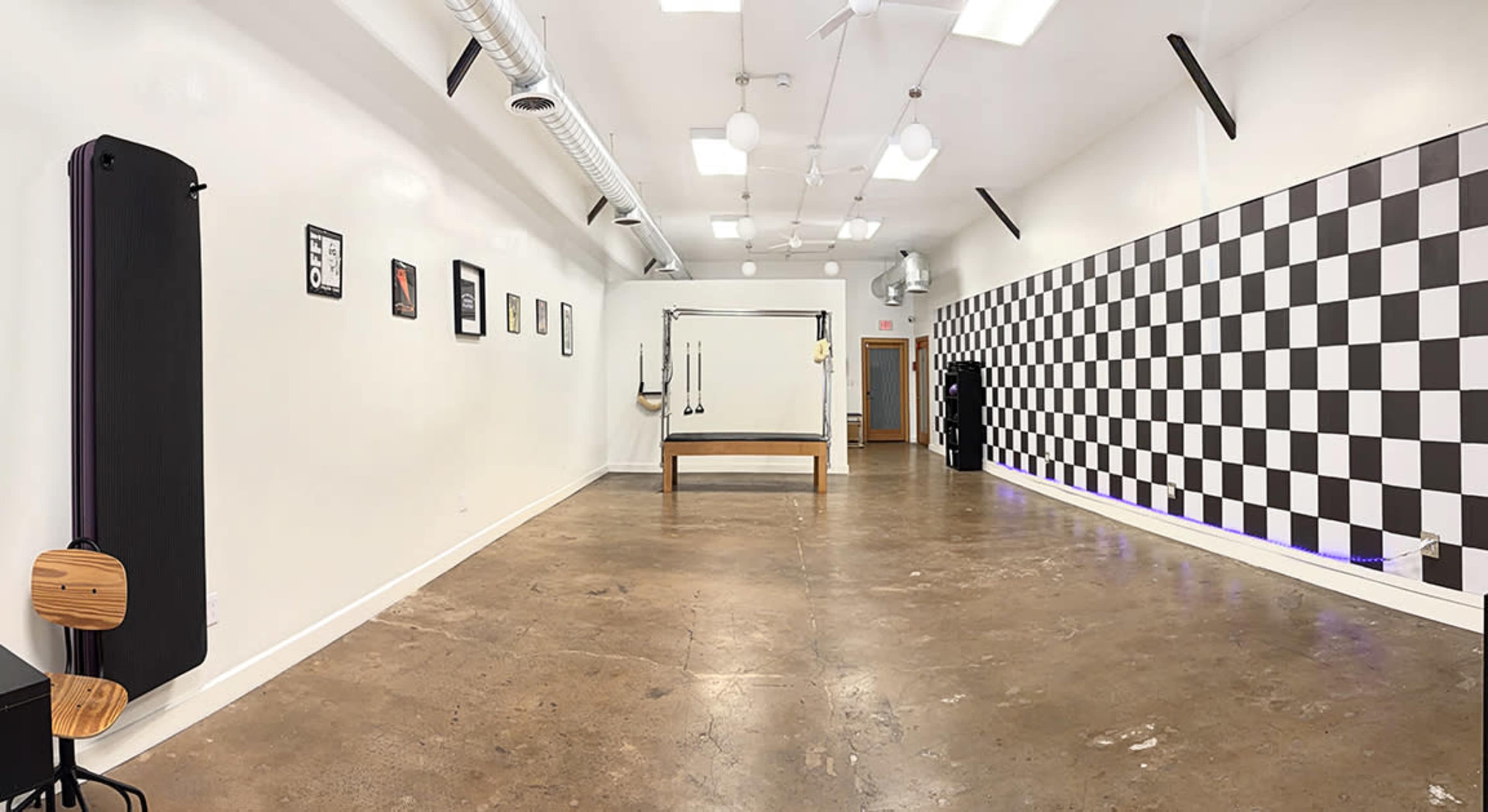 The image shows a spacious, well-lit room with a concrete floor and a black-and-white checkered wall, featuring a table, exercise equipment, and framed art on the walls.