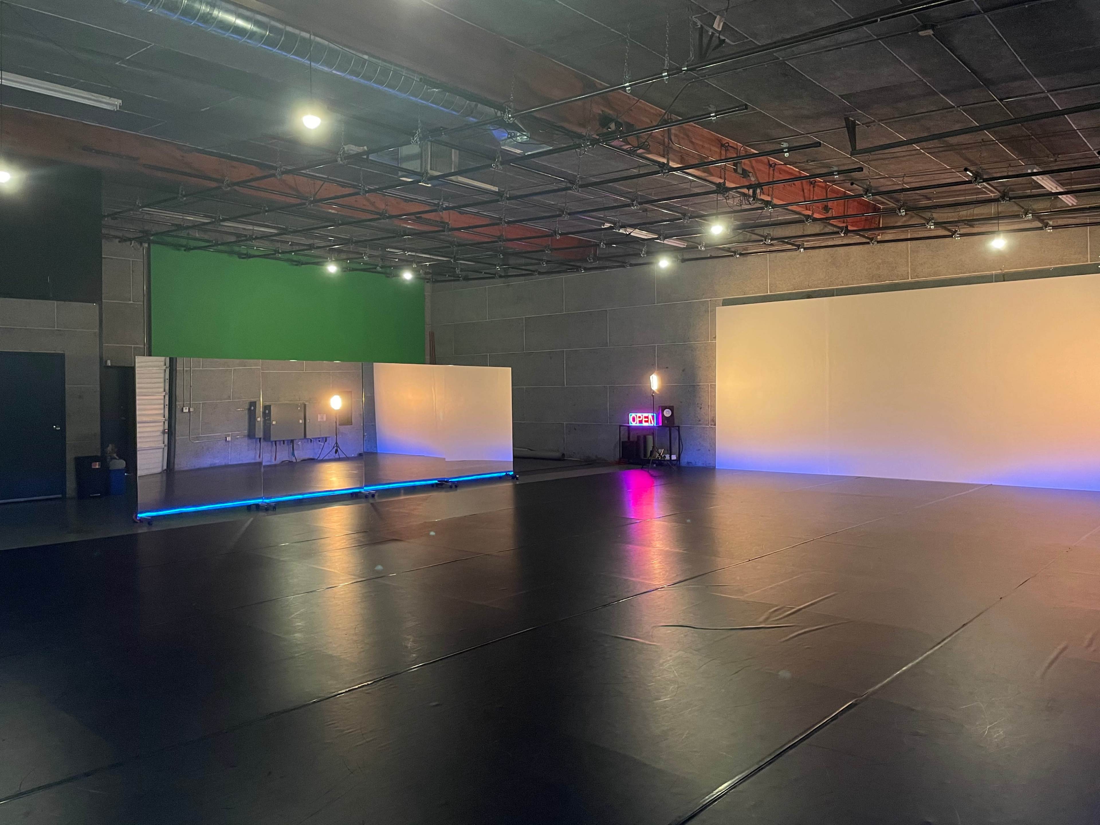 A spacious studio with dark flooring, light walls, and a green screen area, illuminated by several artificial lights.