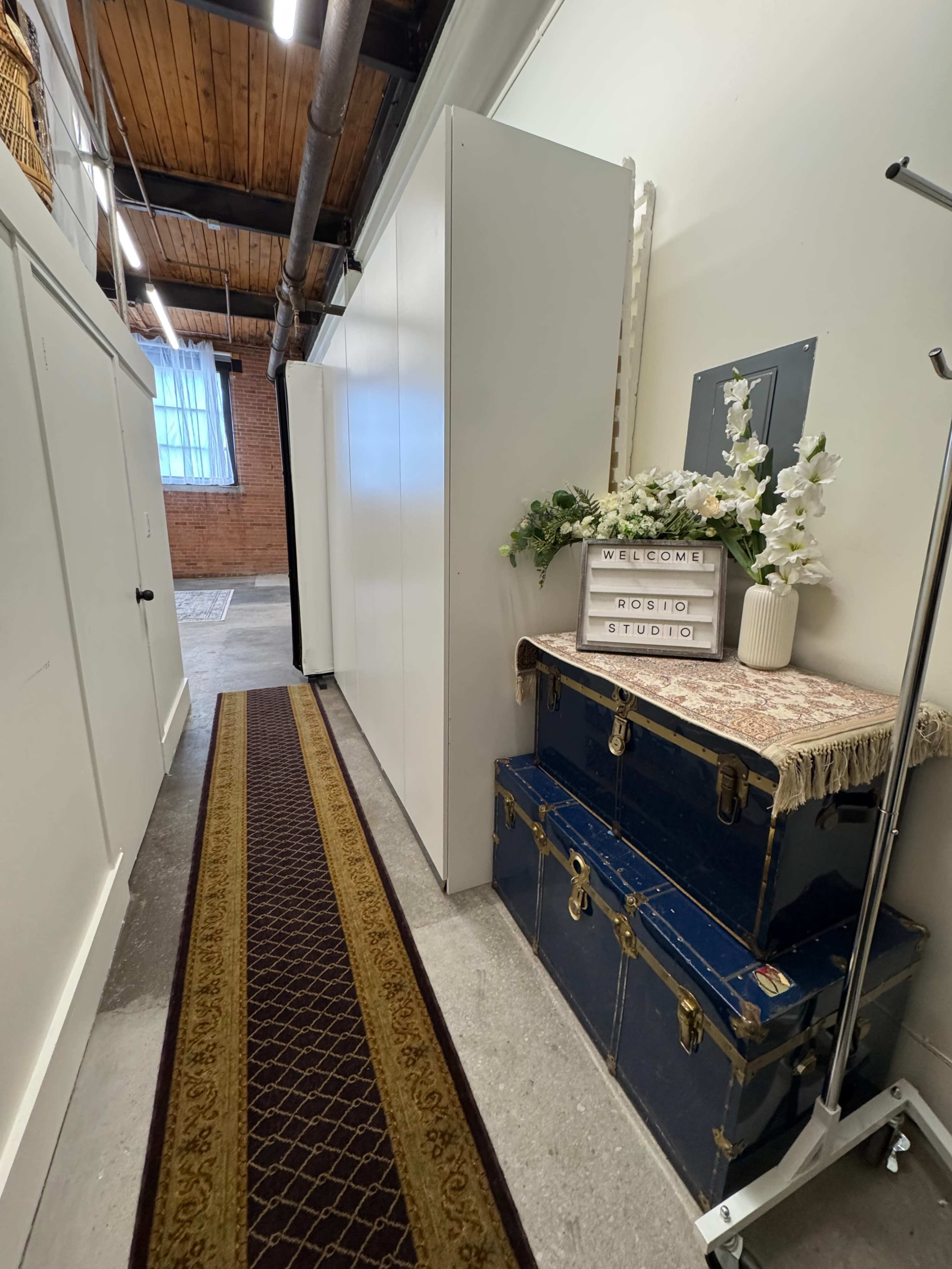 The image shows a narrow hallway with white storage cabinets on one side, a decorative floral arrangement on a wooden table, and a blue trunk at the bottom.