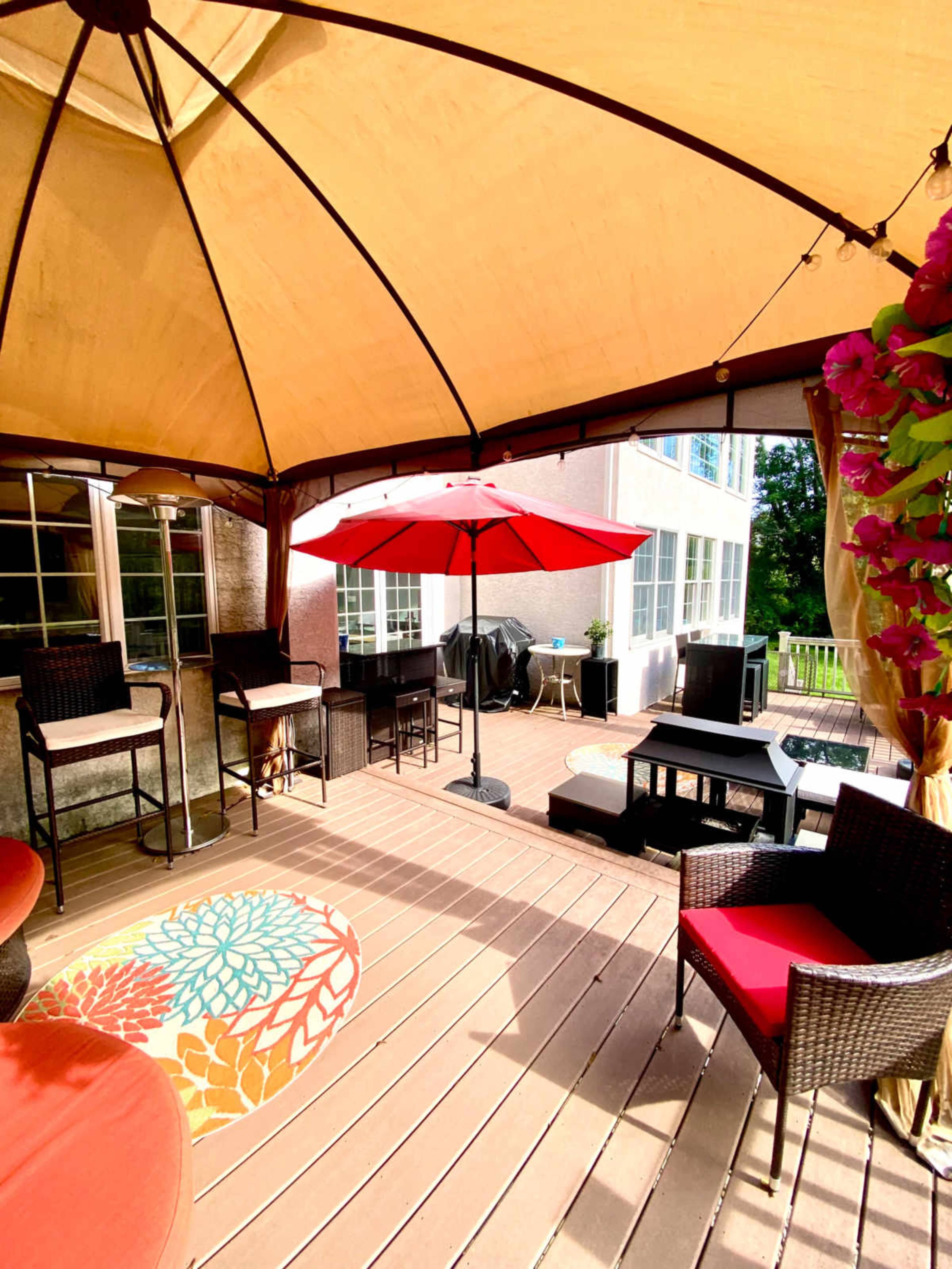 A shaded outdoor patio features a red umbrella over a seating area with black wicker furniture and a decorative rug.
