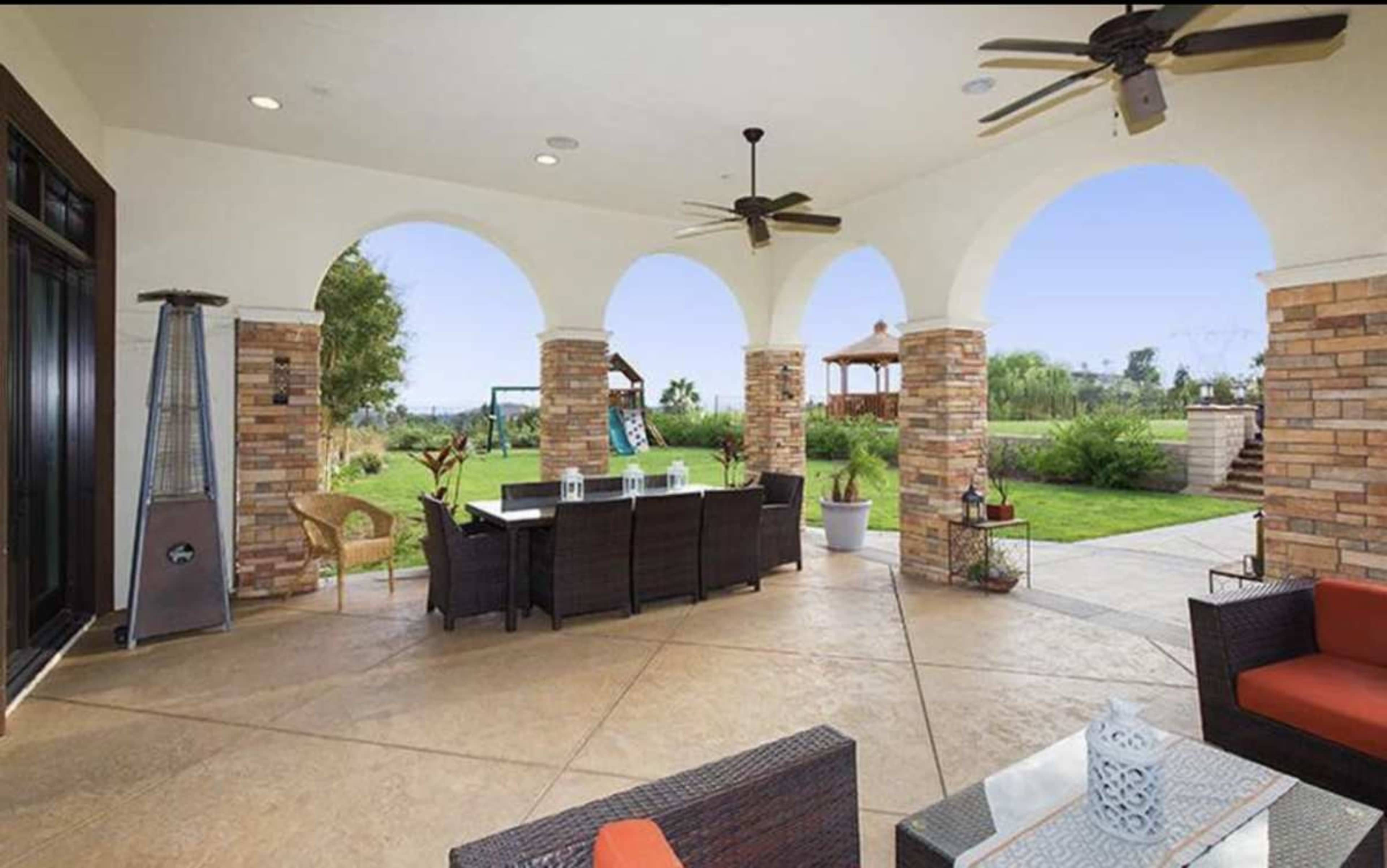 The image shows a covered outdoor patio with a seating area, stone pillars, and a landscaped garden visible in the background.
