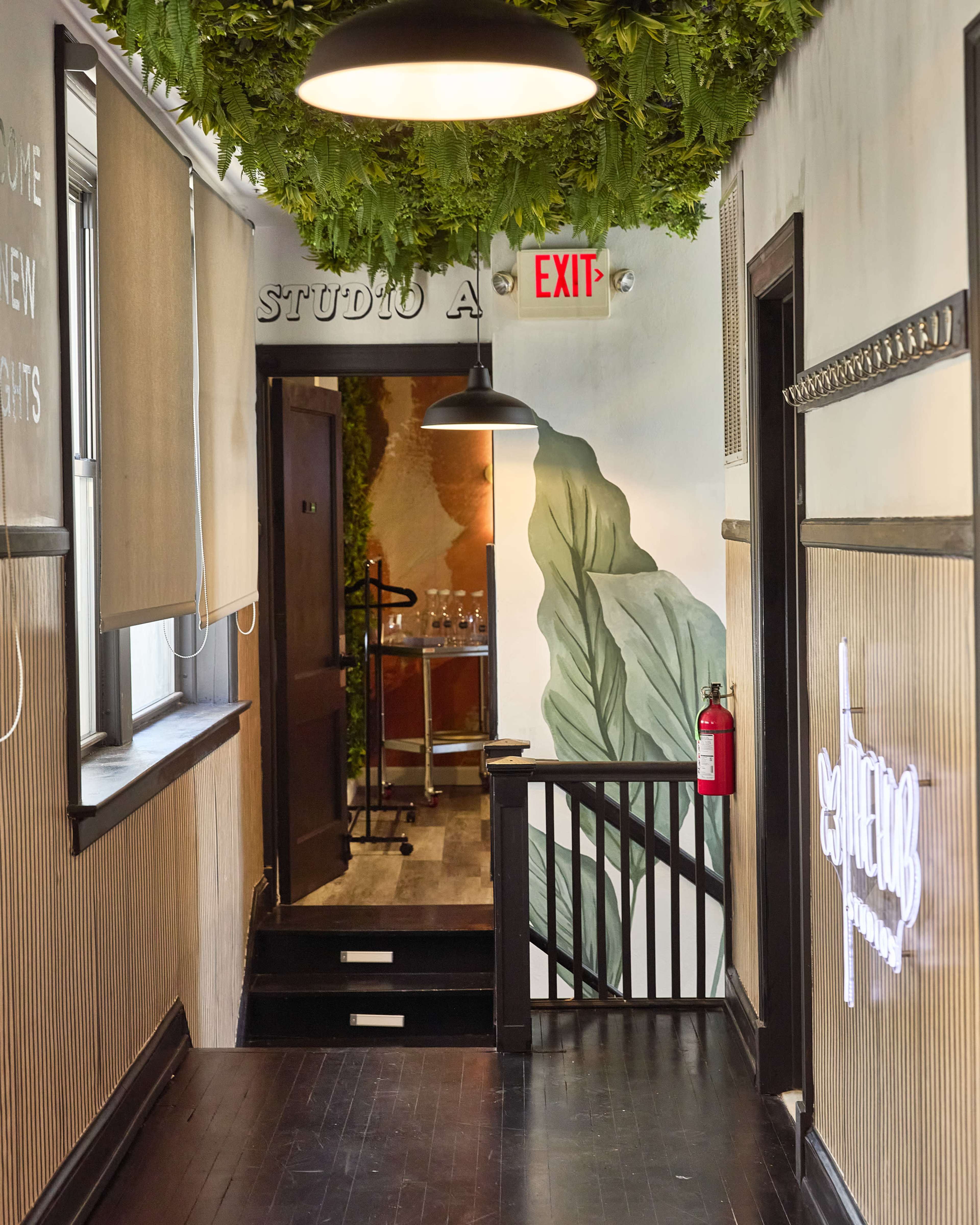 The image shows a hallway adorned with greenery, leading to an exit and featuring a combination of wood paneling and wall art.
