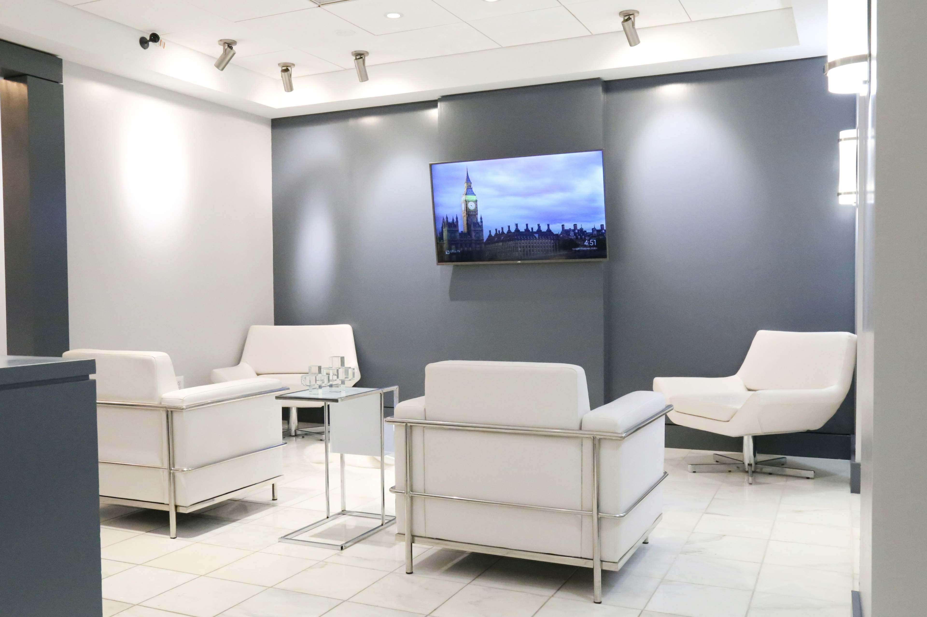 A modern waiting area features three white chairs, a glass table, and a television displaying an image of Big Ben.