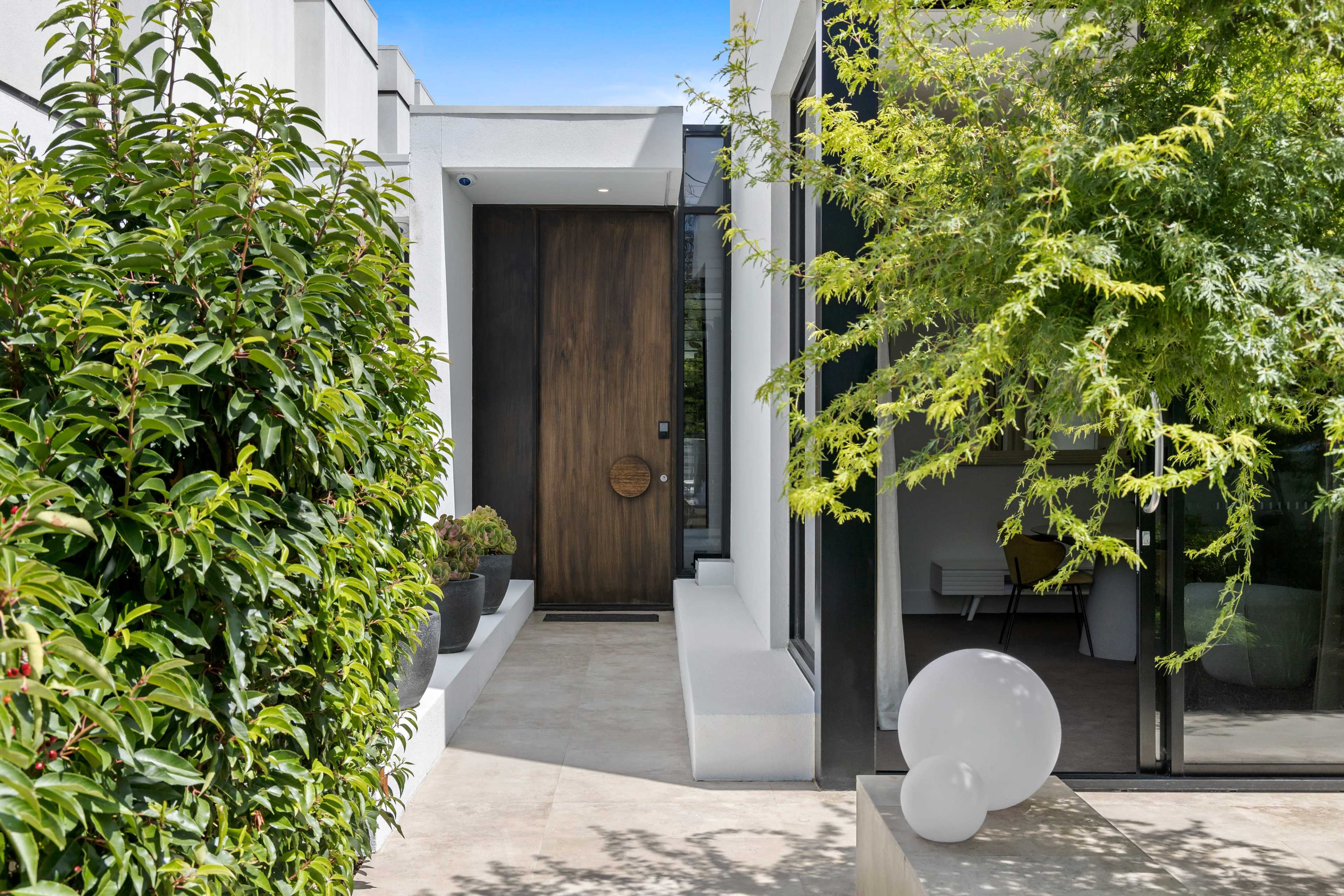 A modern entrance with a wooden door surrounded by greenery and a glass wall showcasing an interior space.