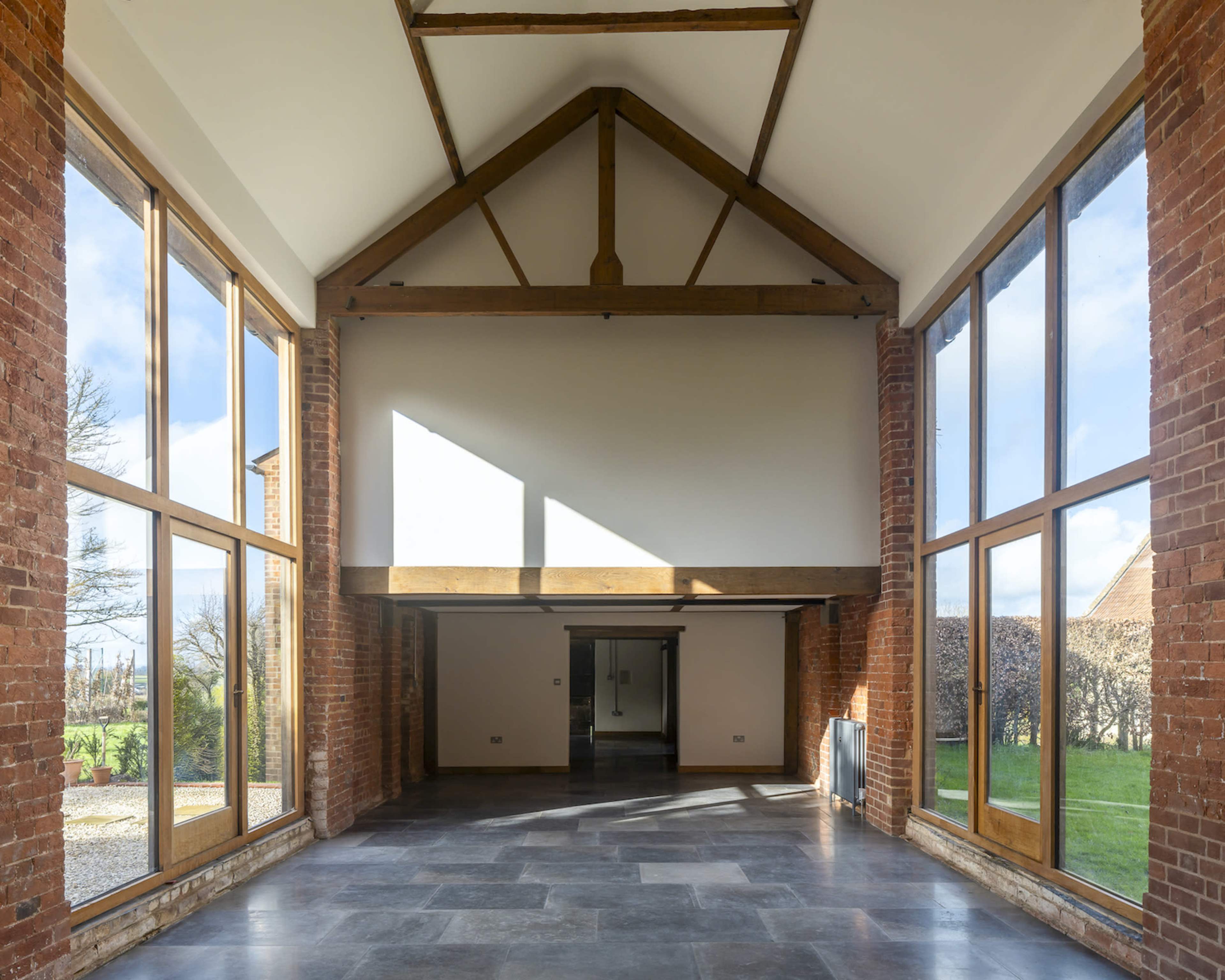 The image shows a large, open interior space with high ceilings, characterized by brick walls and expansive windows allowing natural light to flood in.
