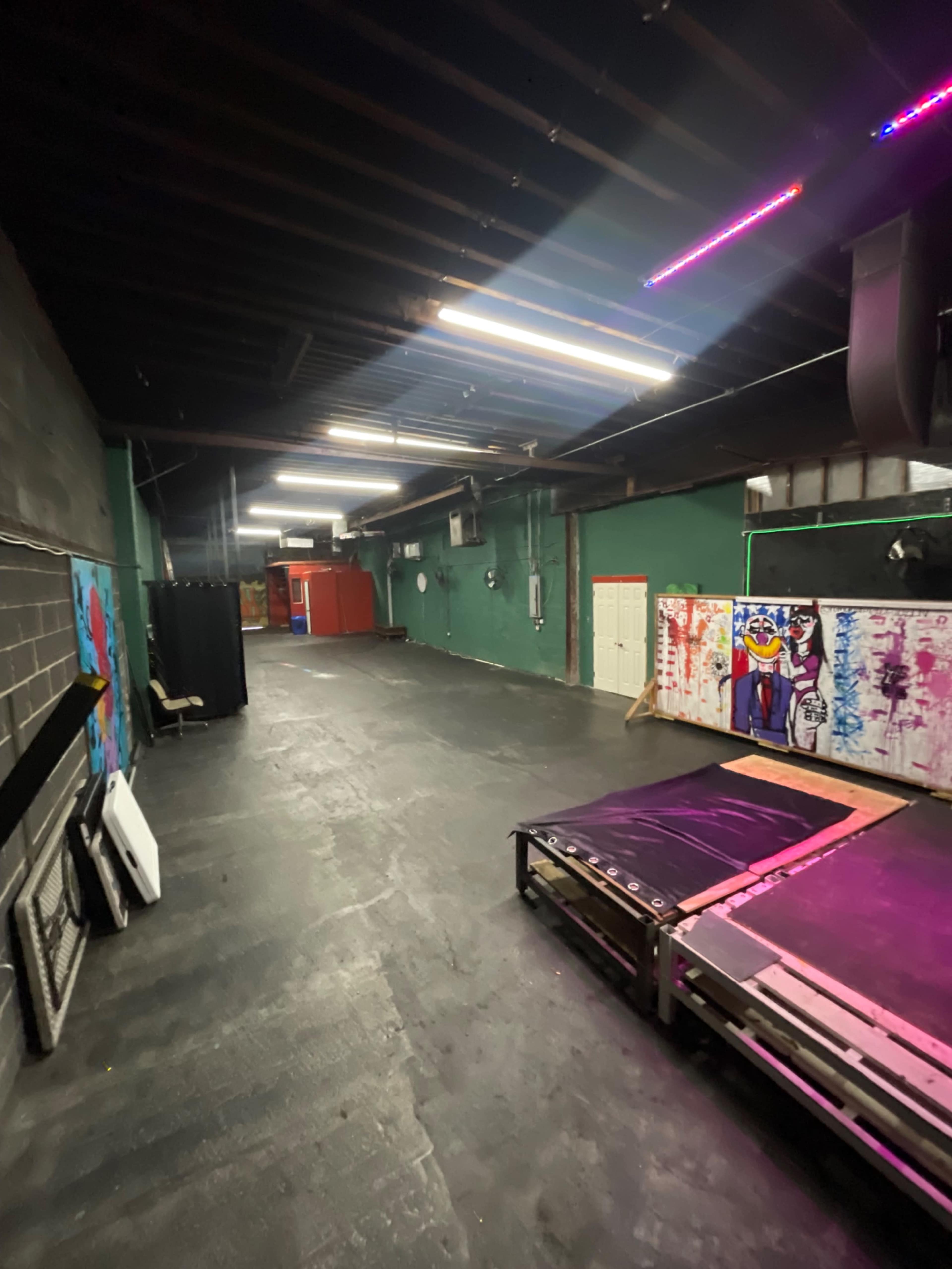 The image shows an empty industrial-style room with dark flooring, green walls, and various colorful murals on one side, illuminated by overhead lights.