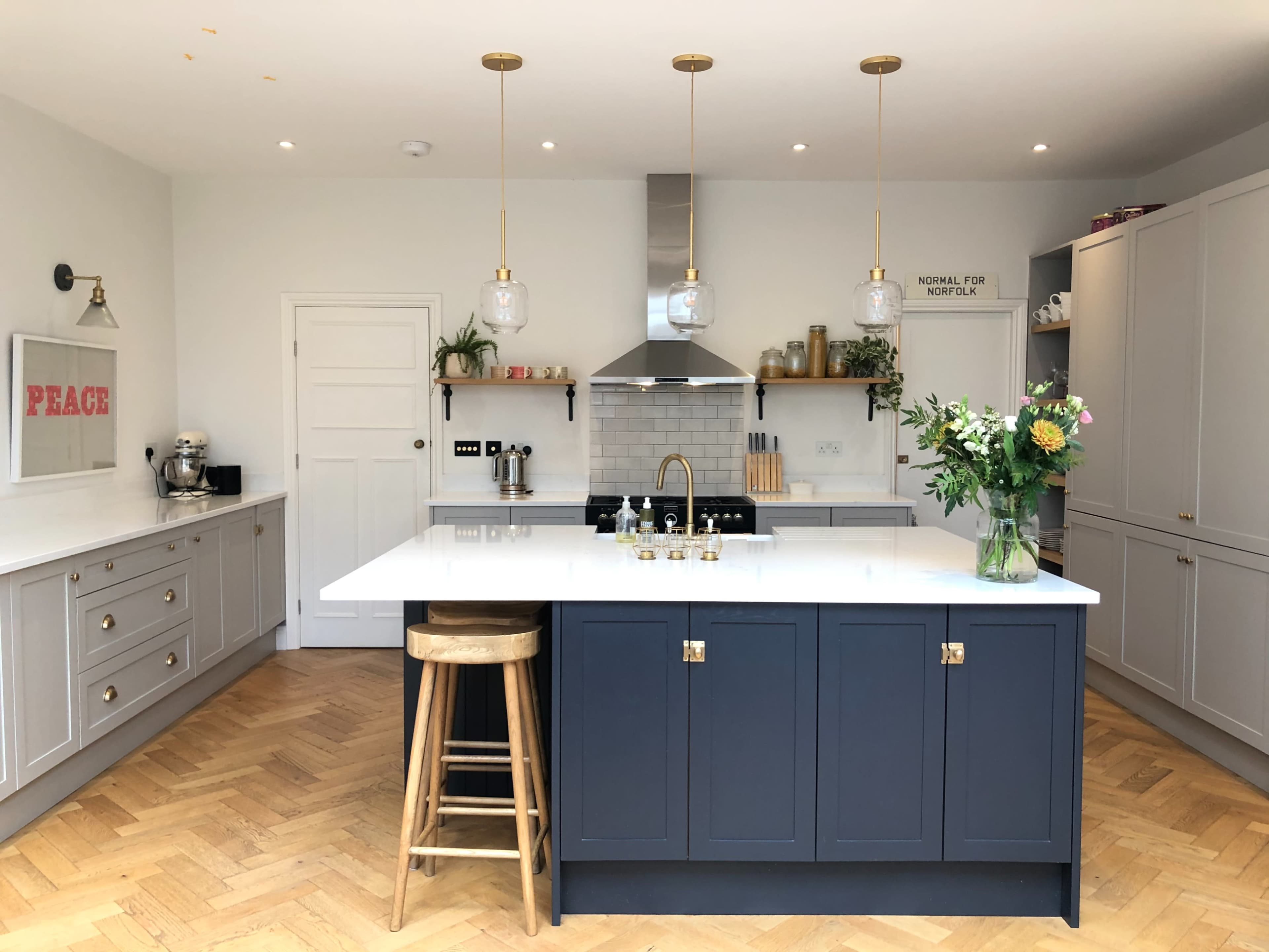 A modern kitchen features a central island with a dark blue base, white countertop, and brass fixtures, surrounded by light cabinetry and wooden flooring.