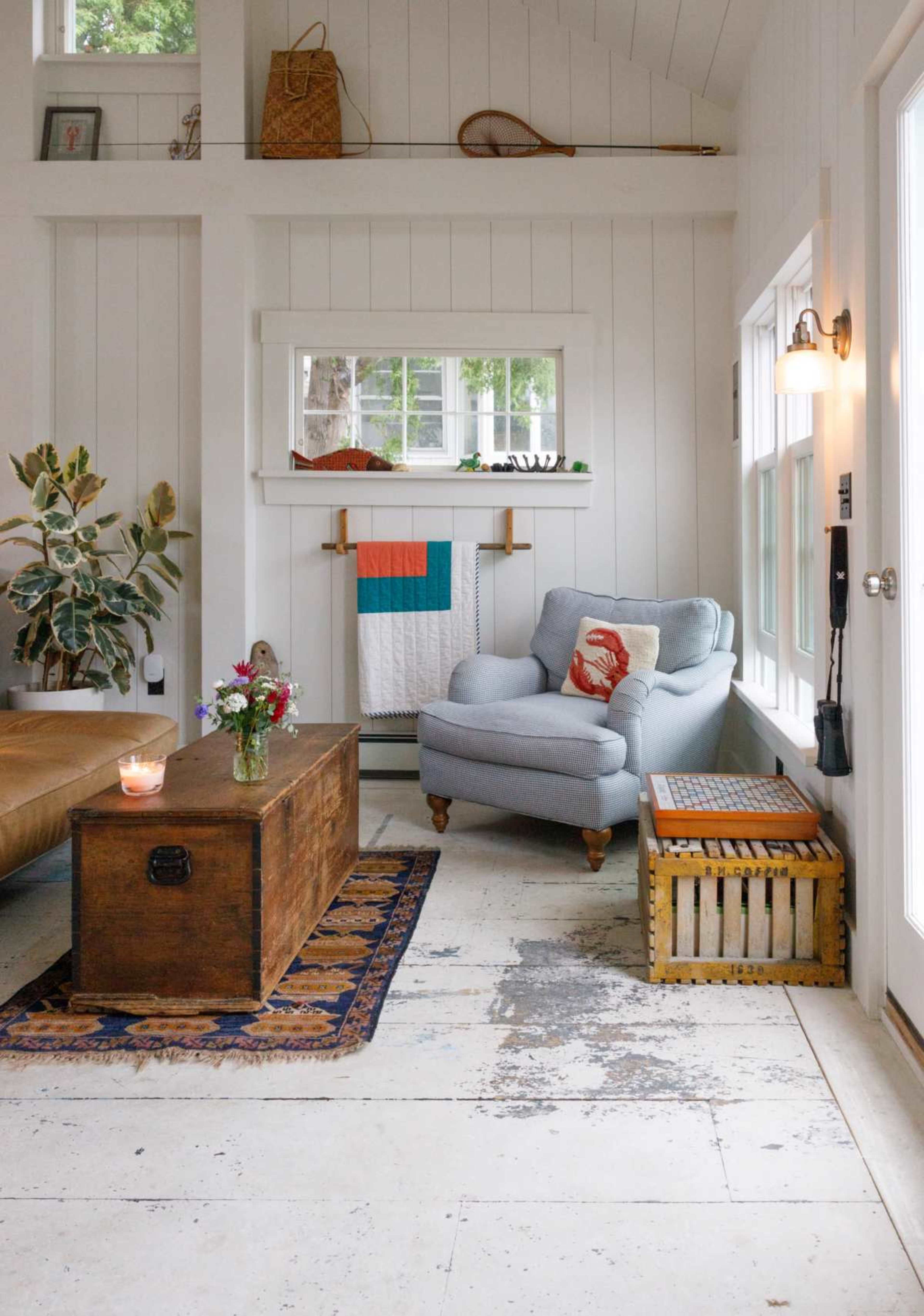 The image shows a cozy living room with a light blue armchair, a wooden coffee table, and a quilt displayed on the wall, surrounded by white paneling and large windows.