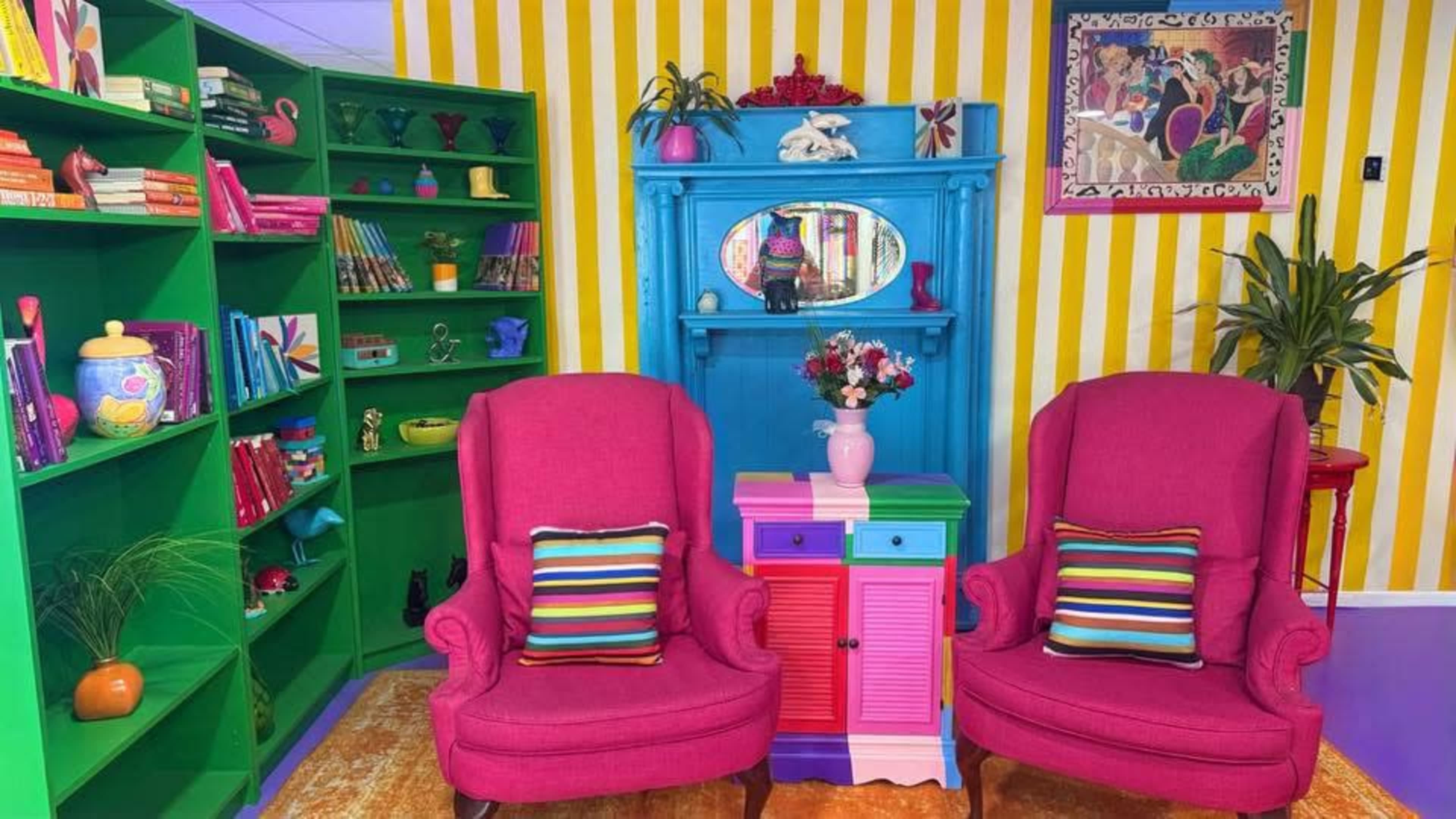 A vibrant room features two pink armchairs with striped cushions situated in front of a colorful bookshelf and a blue cabinet, all set against yellow striped walls.