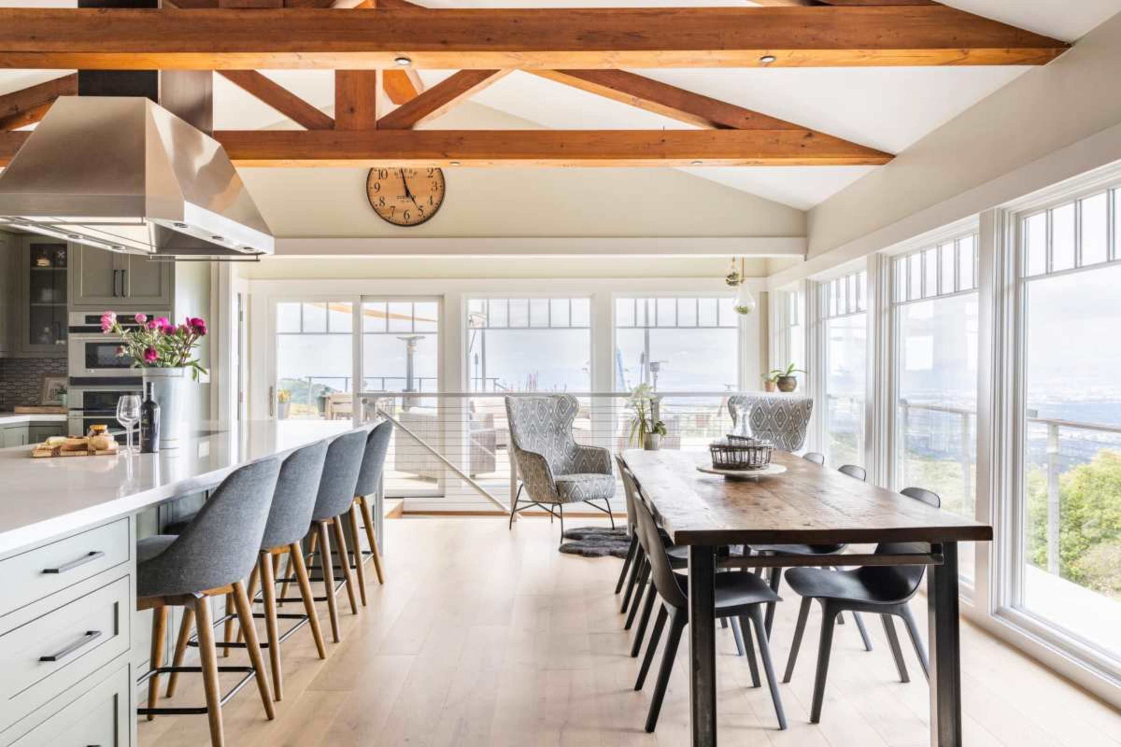A modern kitchen and dining area features a large island with barstools, a wooden dining table, and large windows offering a view of the outdoors.
