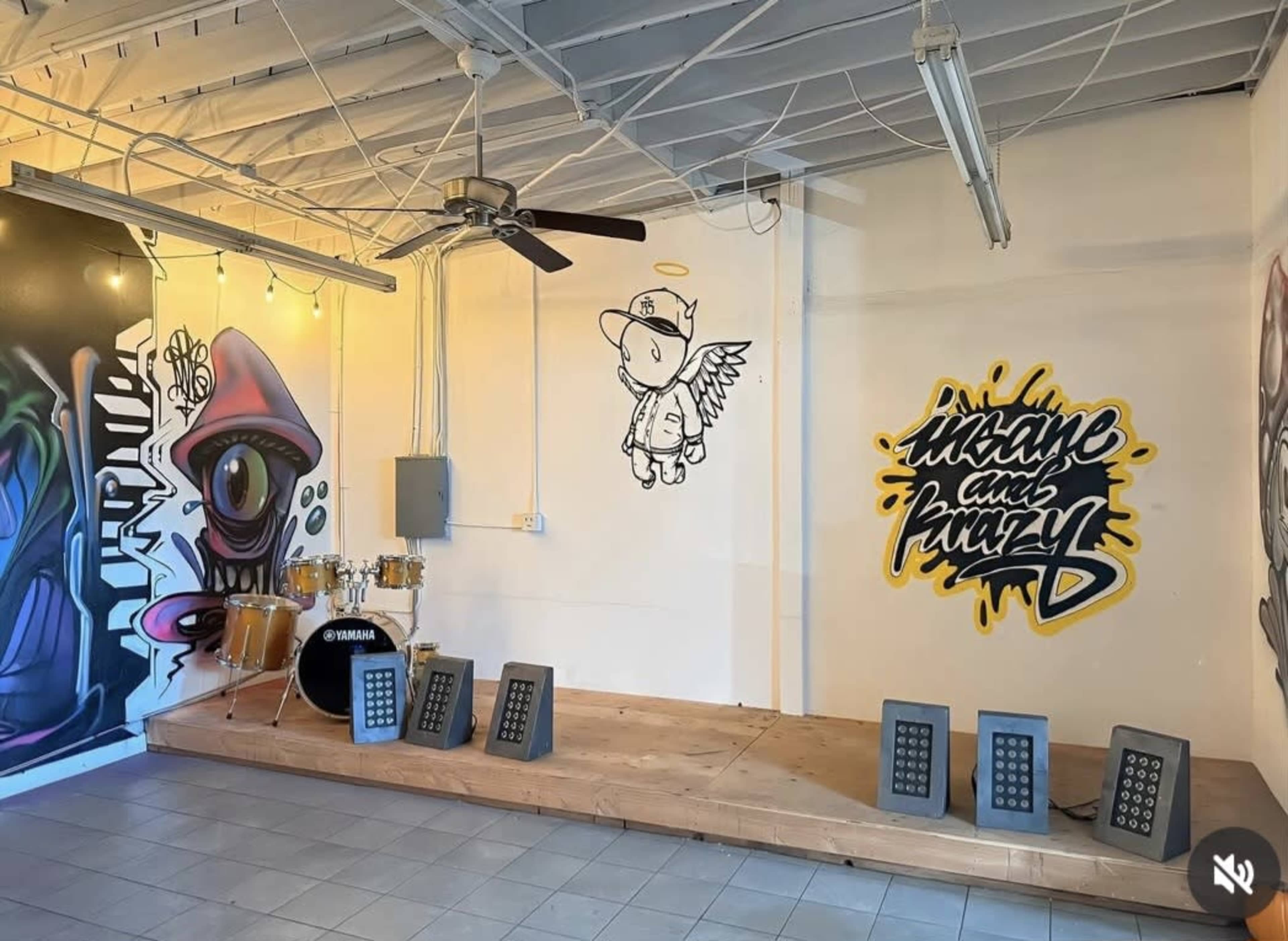 The image shows an interior space featuring a wooden platform with speakers, a drum set, and graffiti art on the walls.