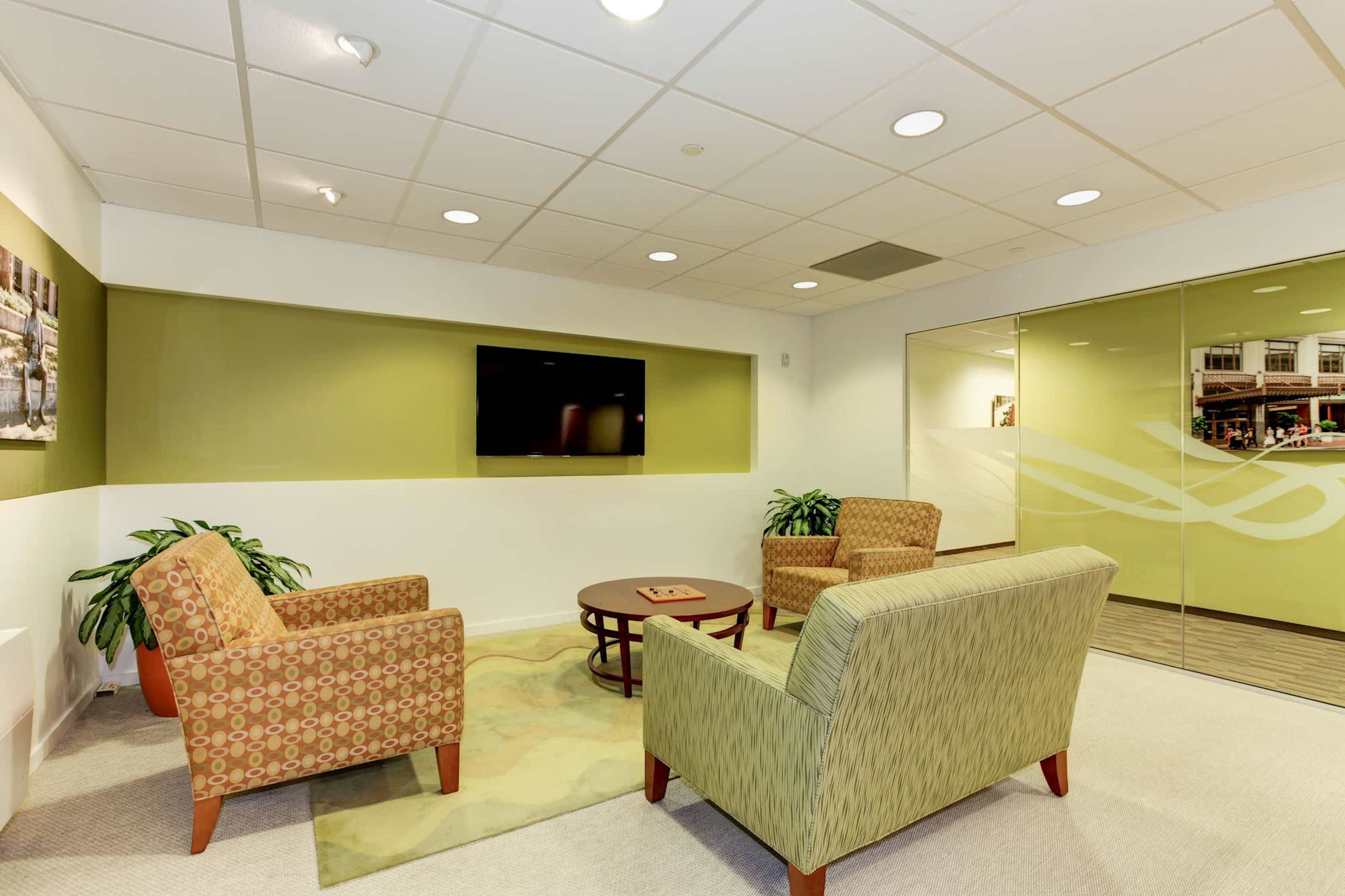 A modern office waiting area with two patterned chairs, a round table, and a wall-mounted television surrounded by light green walls.