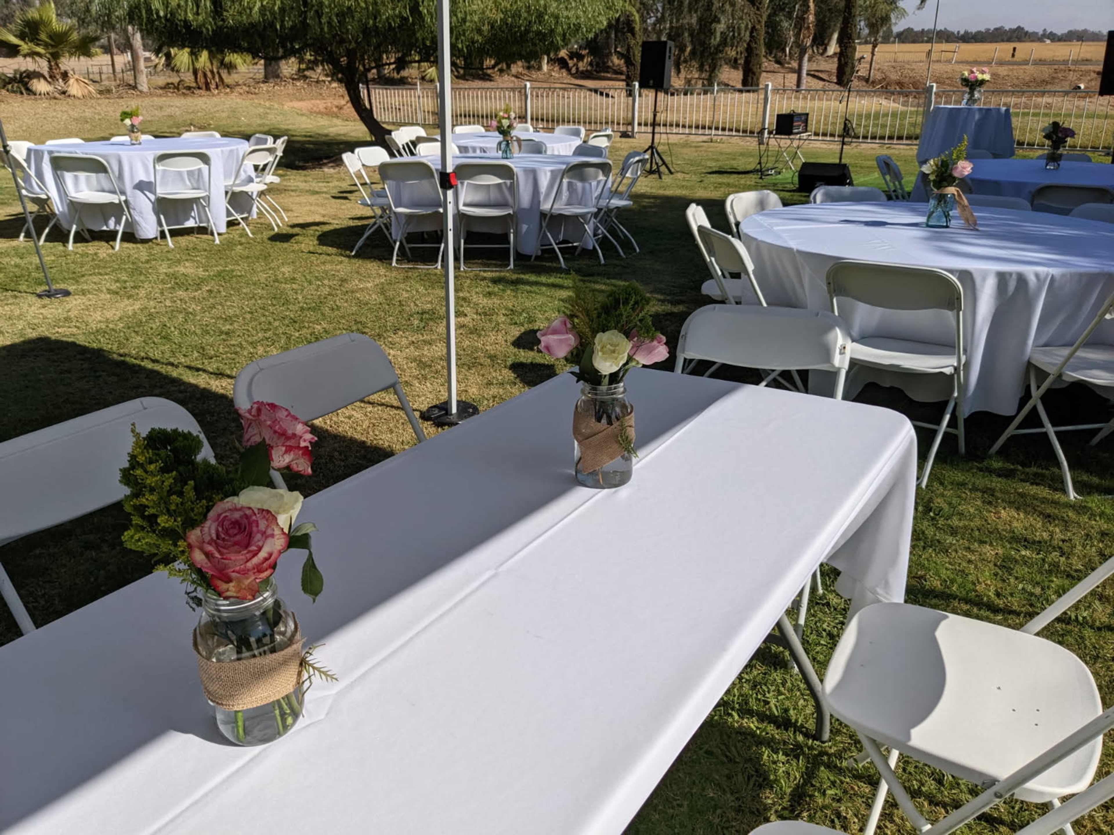 The image shows a set of tables with white tablecloths and mason jars filled with flowers, set up in a grassy outdoor area for an event.