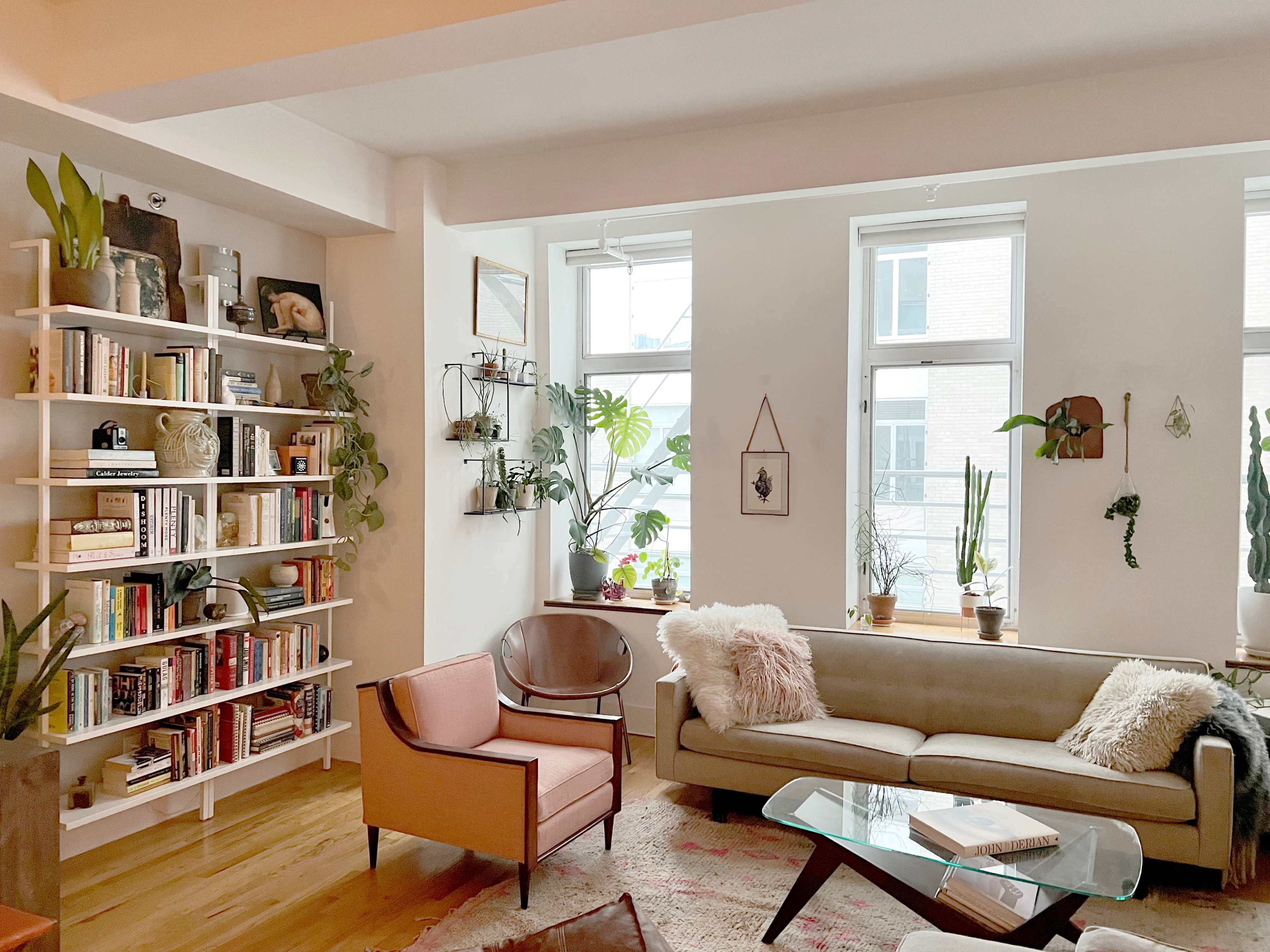 A cozy living room with a bookshelf filled with books, a comfortable sofa, an armchair, and various indoor plants arranged by large windows.
