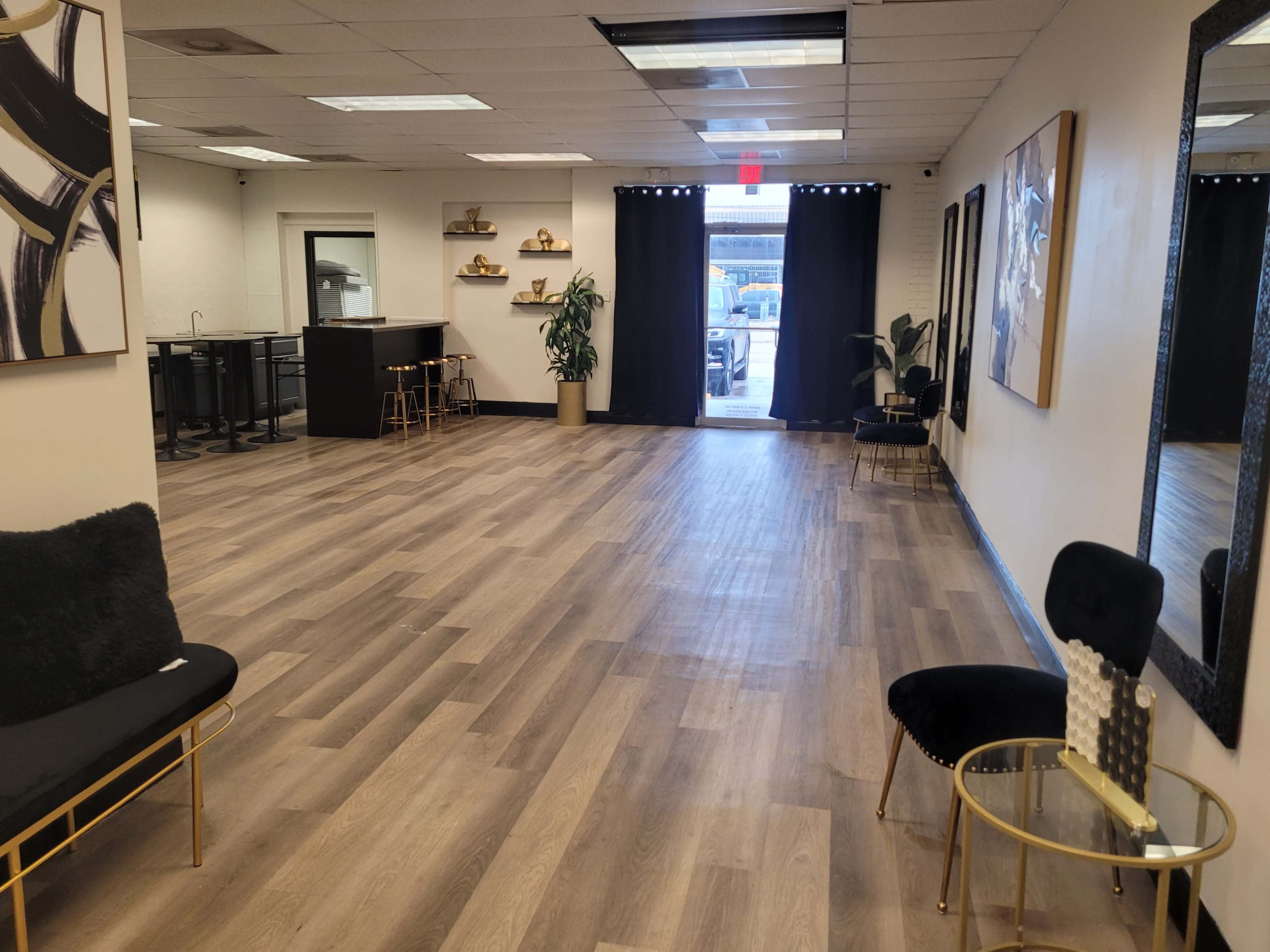 The image shows a spacious interior room with wooden flooring, featuring a table and chairs along one wall, a plant, and an open door leading outside.