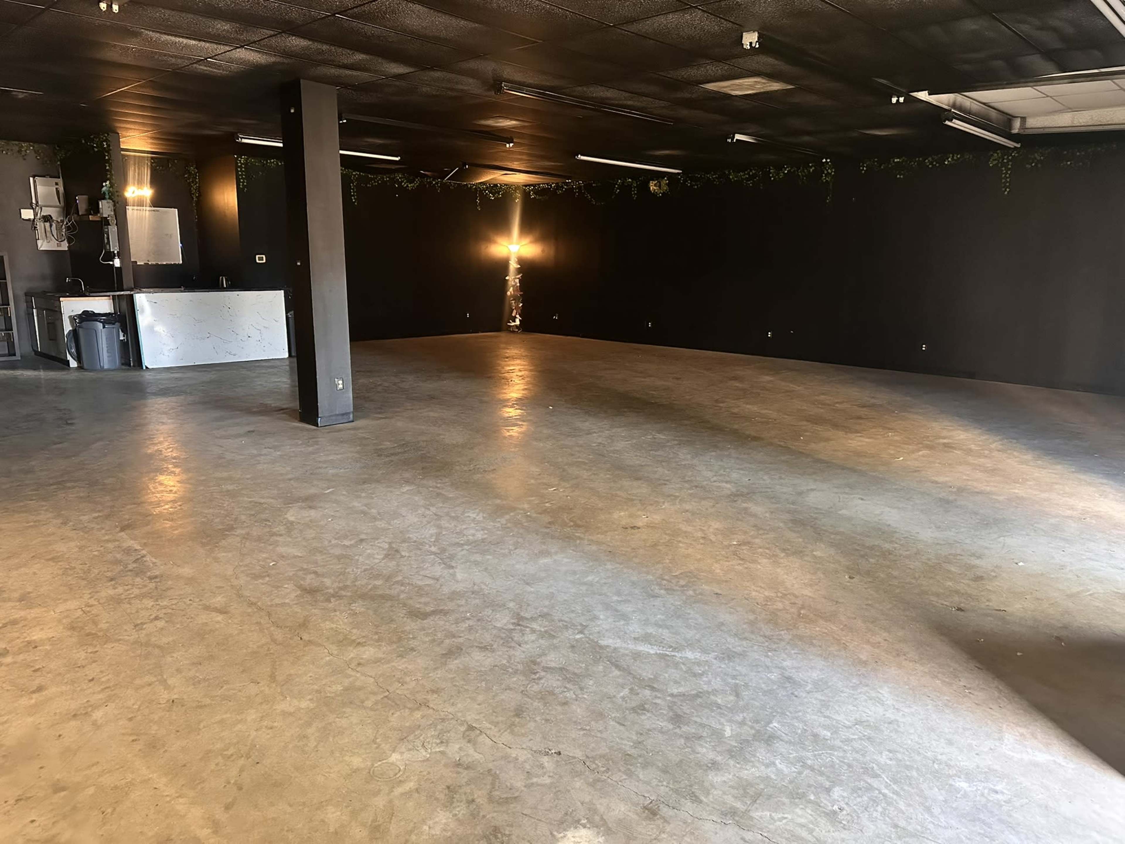 The image shows a spacious, empty room with a polished concrete floor, black walls, and a small bar area in the corner.