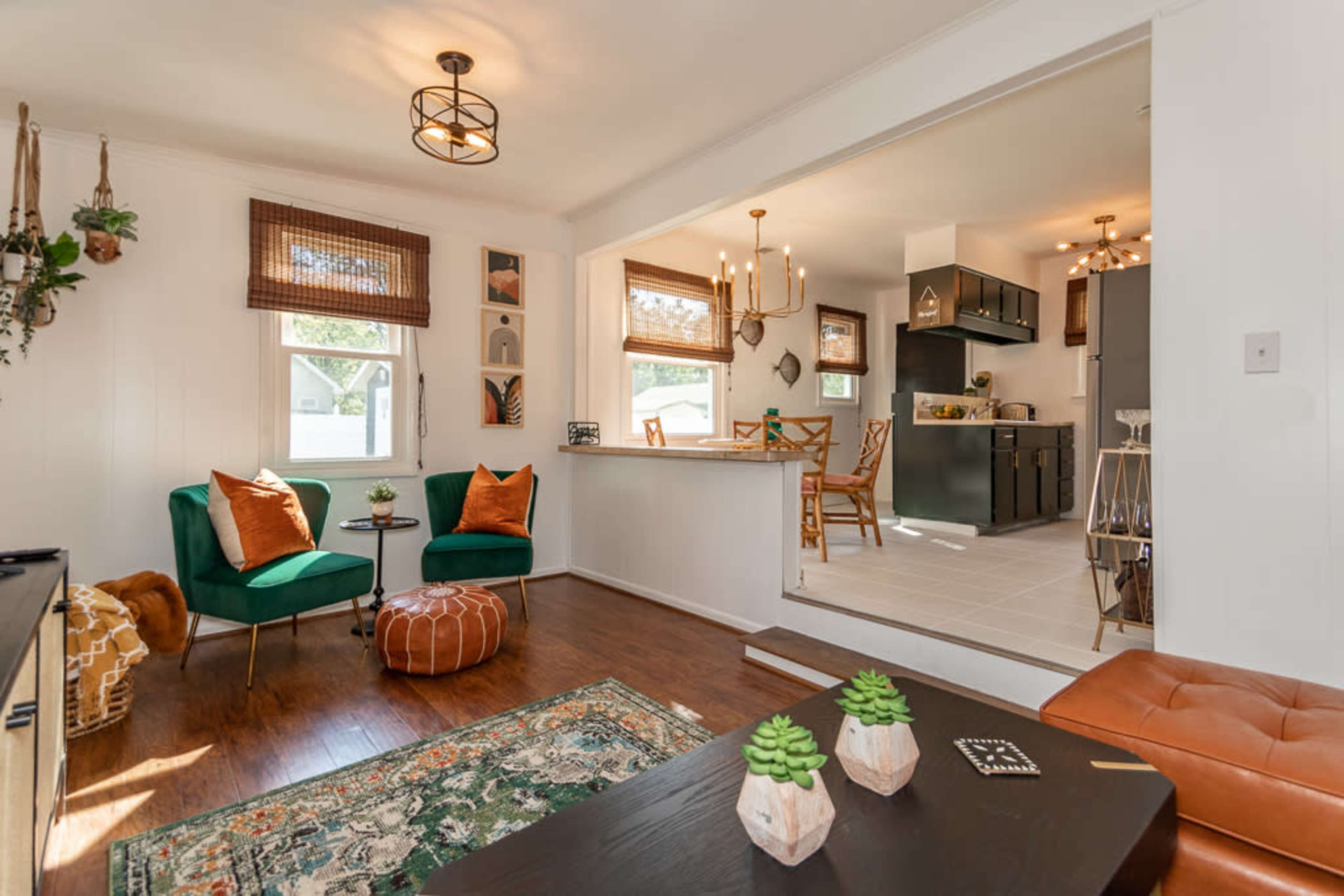 A cozy living area with two green chairs, a round ottoman, and a view into a kitchen featuring a dining area.
