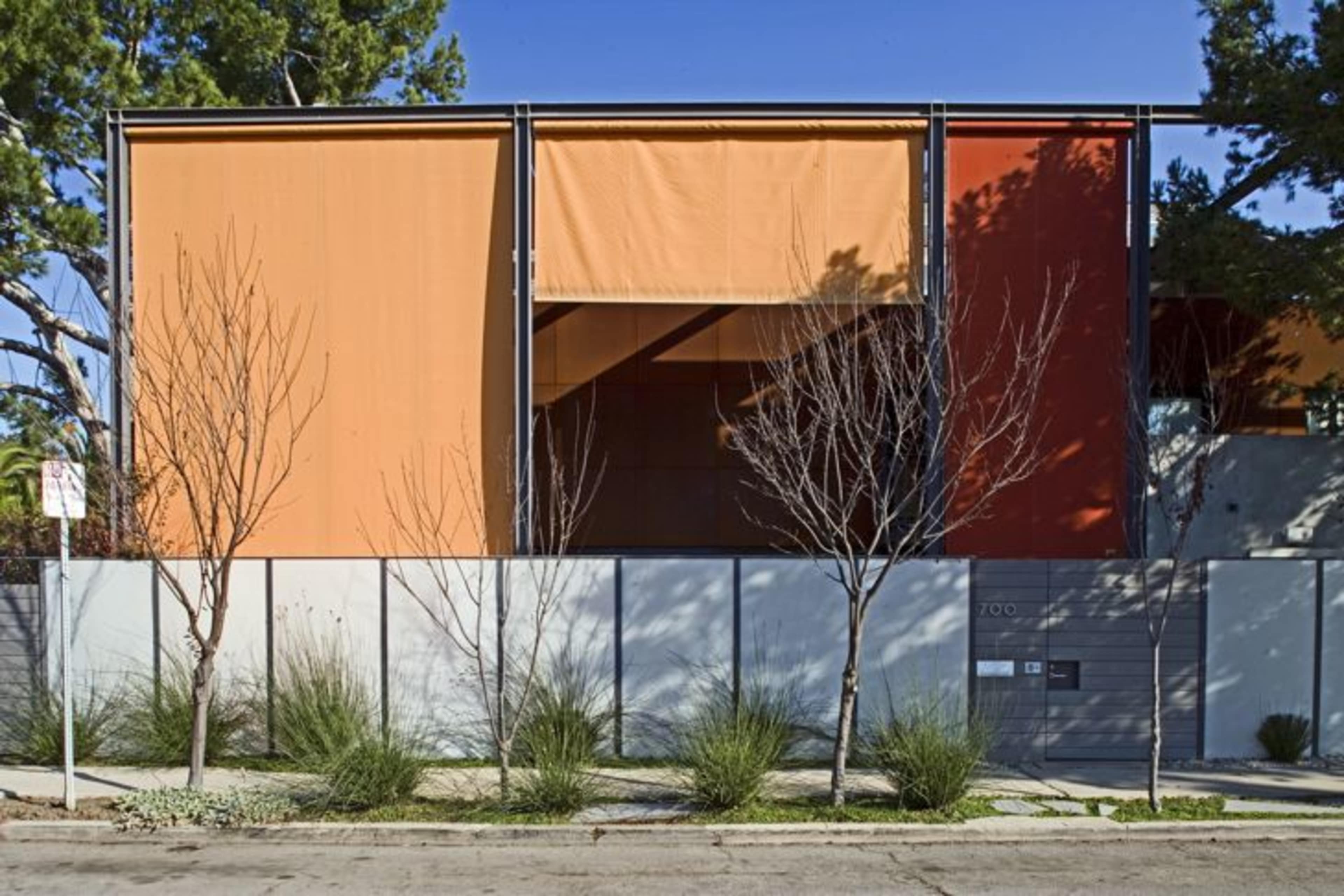 A modern building with large orange and red fabric panels, flanked by sparse trees and low shrubs along a sidewalk.