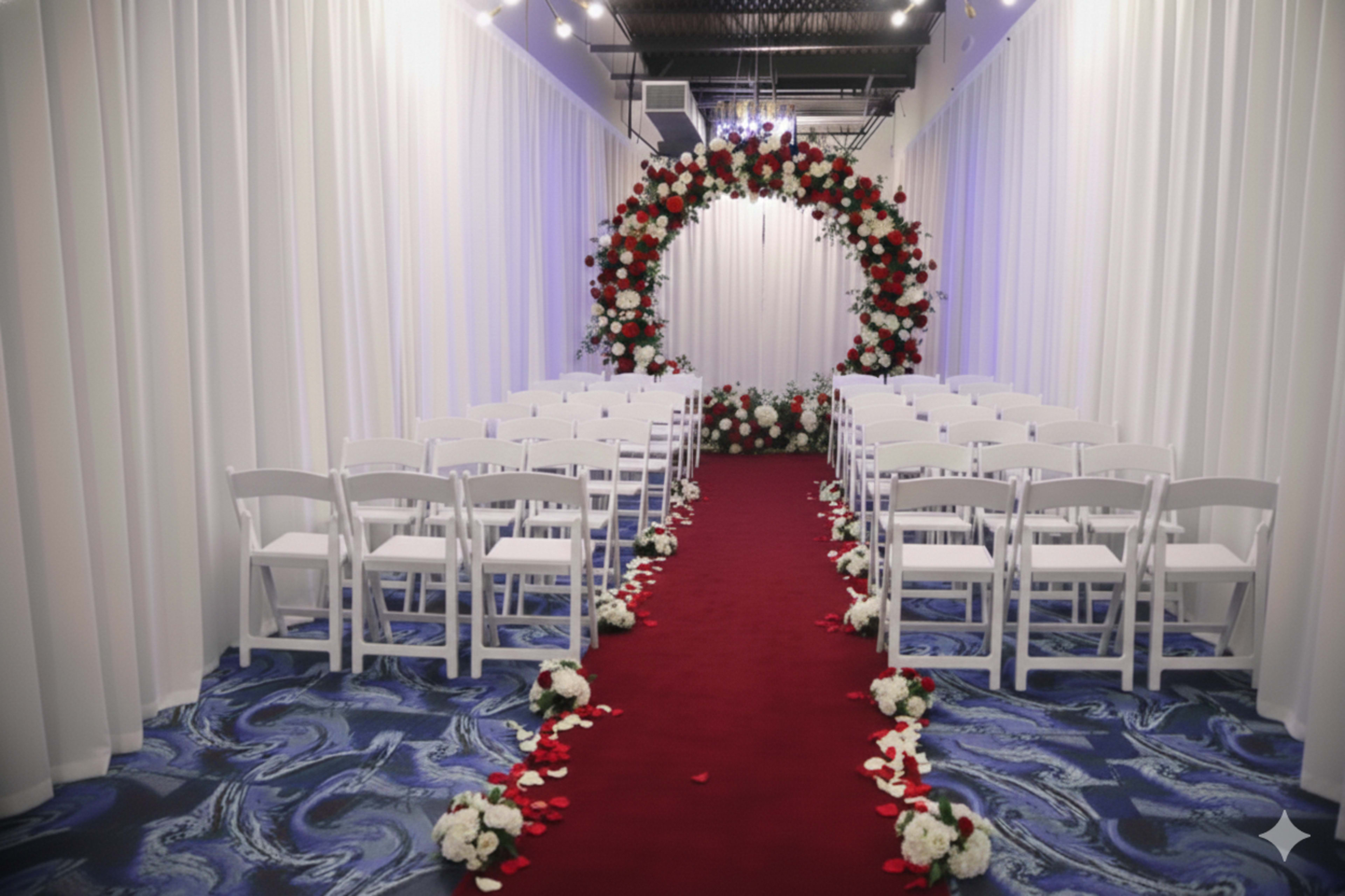 The image shows a wedding ceremony setup with white folding chairs arranged in rows facing a floral arch at the front, adorned with red and white flowers, and a red carpet lined with rose petals.