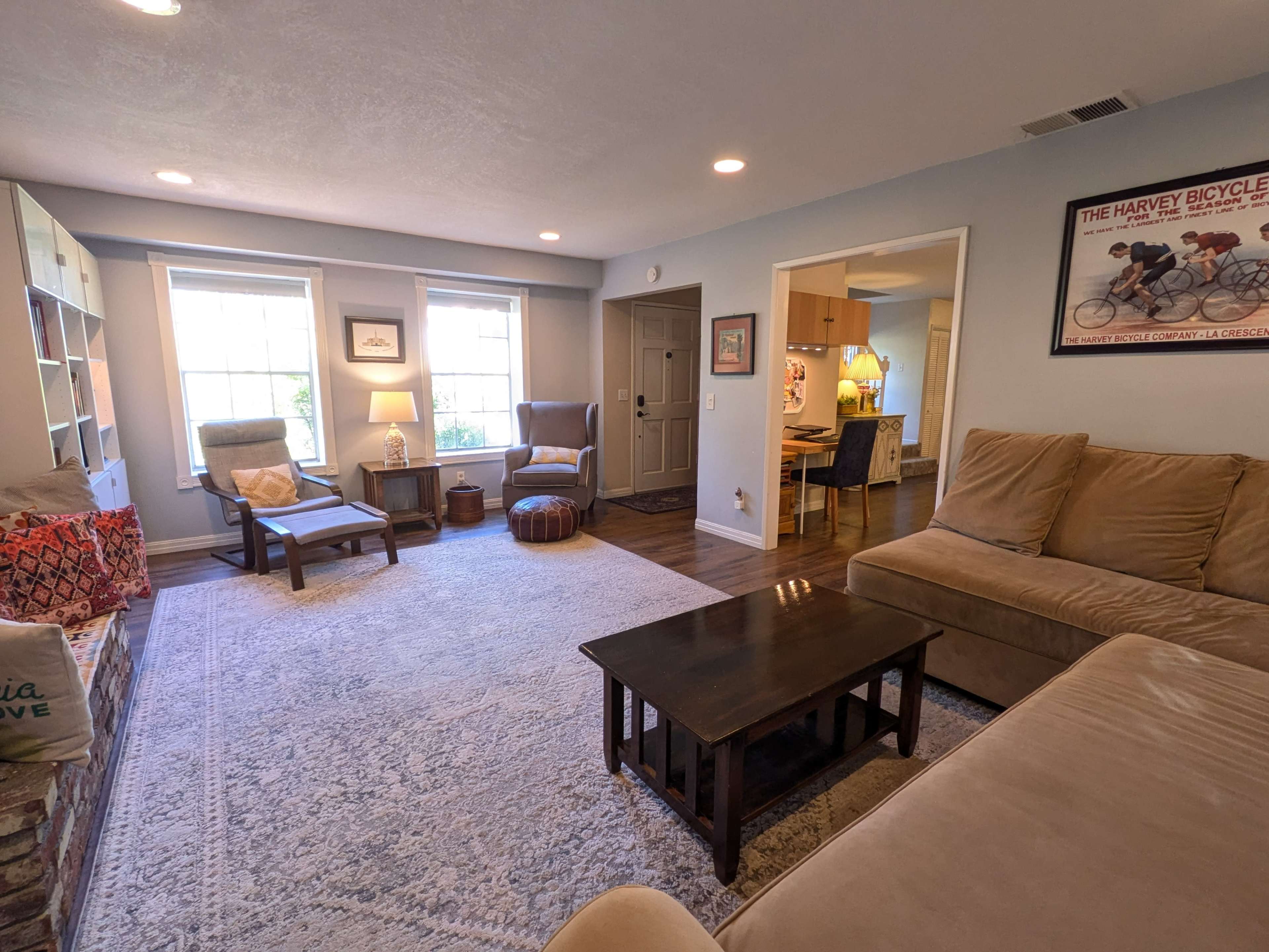 The living room features a light-colored sofa, a dark wooden coffee table, and large windows that illuminate a cozy arrangement of chairs and decorative elements.