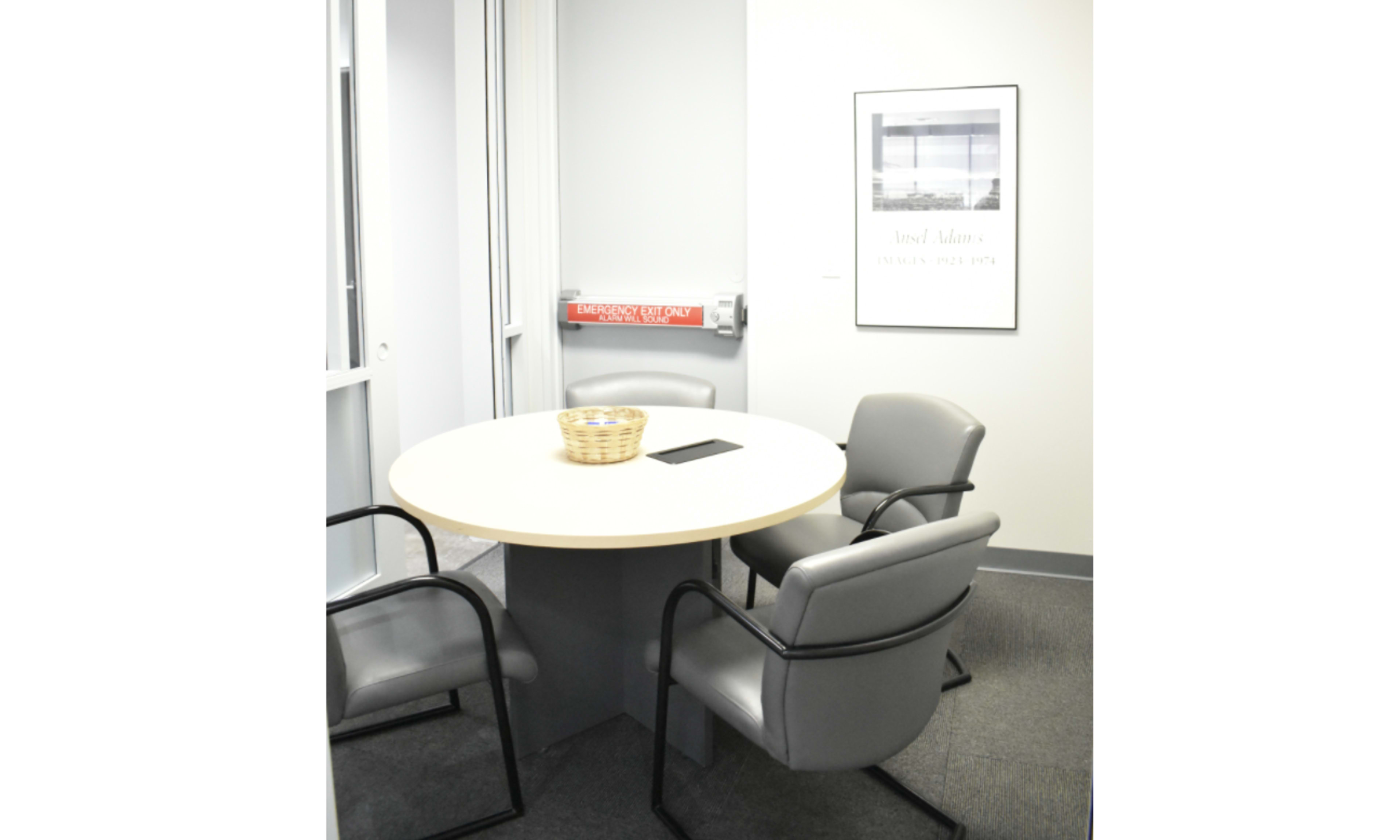A round table with three gray chairs and a basket sits in a small office meeting room, featuring a framed picture on the wall and an emergency exit sign.