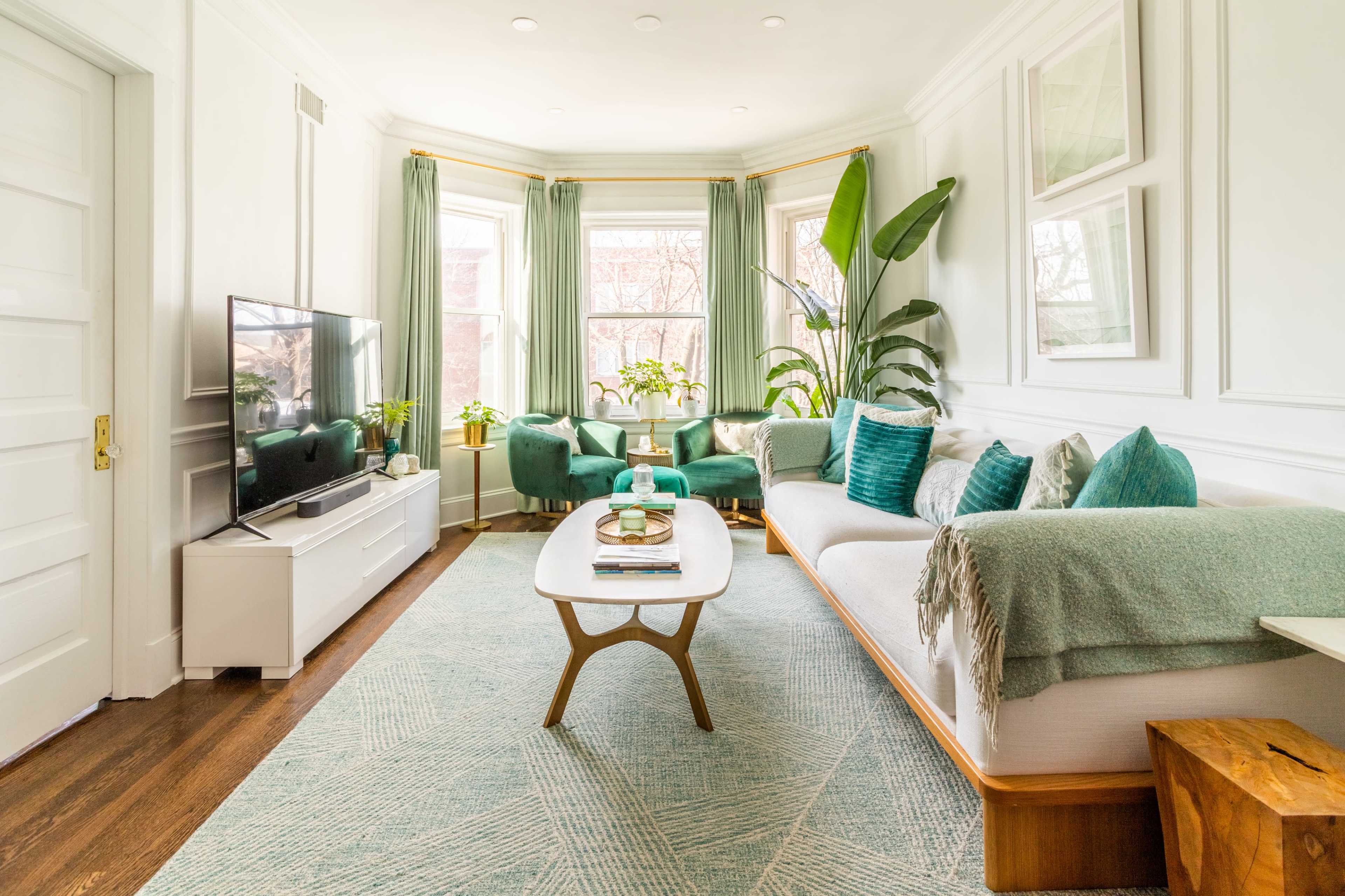 The living room features a modern TV setup, a light-colored sofa with cushions, a wooden coffee table, and large windows with plants, all framed by soft green curtains.